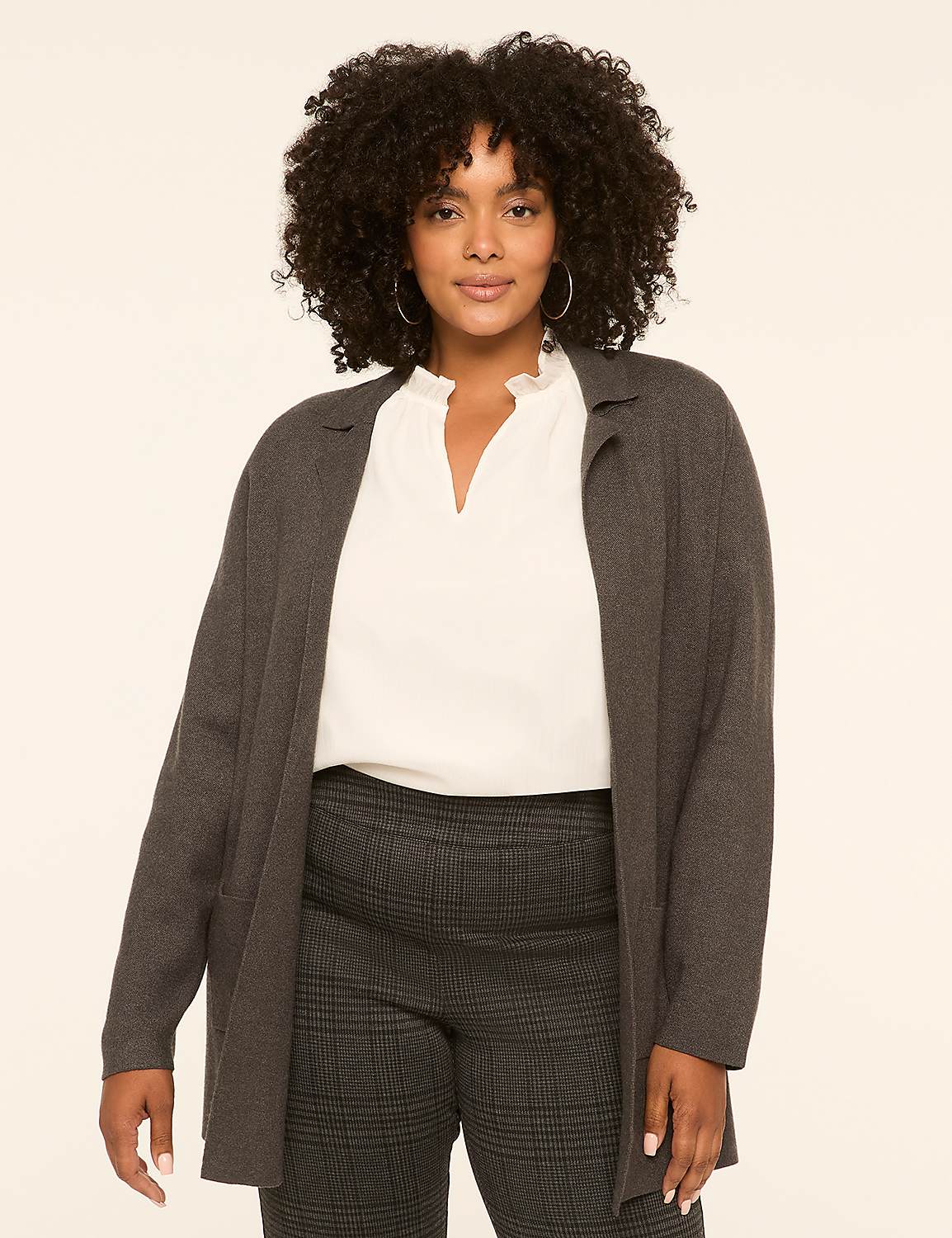A woman with curly dark hair stands in the foreground wearing a gray cardigan over a white blouse and plaid pants, against a plain, light-colored background. Product Image 1