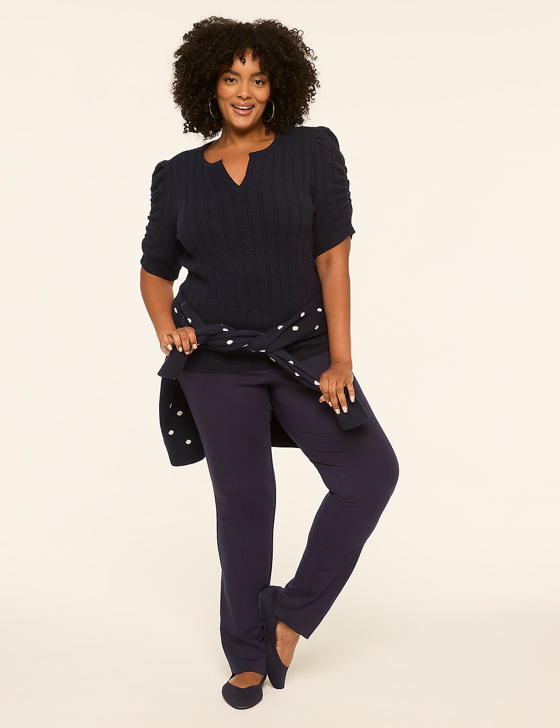 A smiling woman in a navy top and pants stands against a plain, light-colored background, with a polka-dotted sweater tied around her waist. Product Image 3