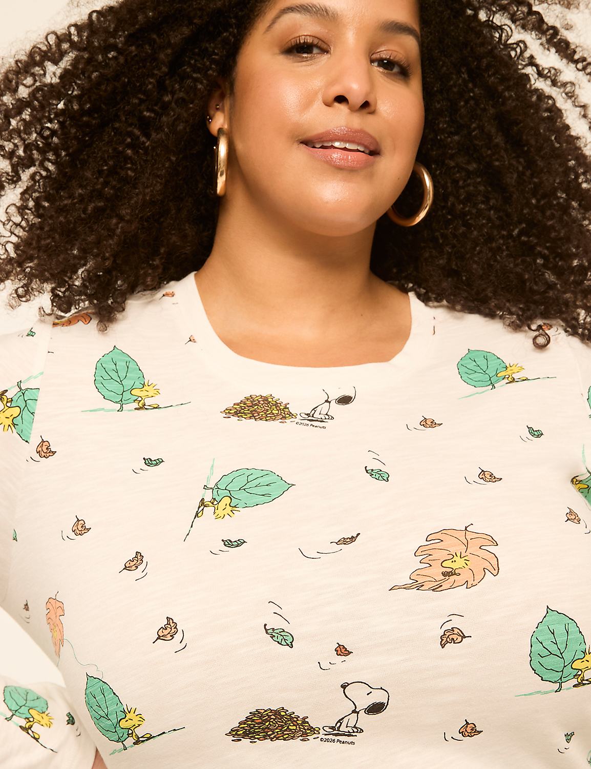 A woman with curly hair and hoop earrings smiles as her cream-colored shirt displays a pattern of falling autumn leaves and Snoopy. Product Image 4