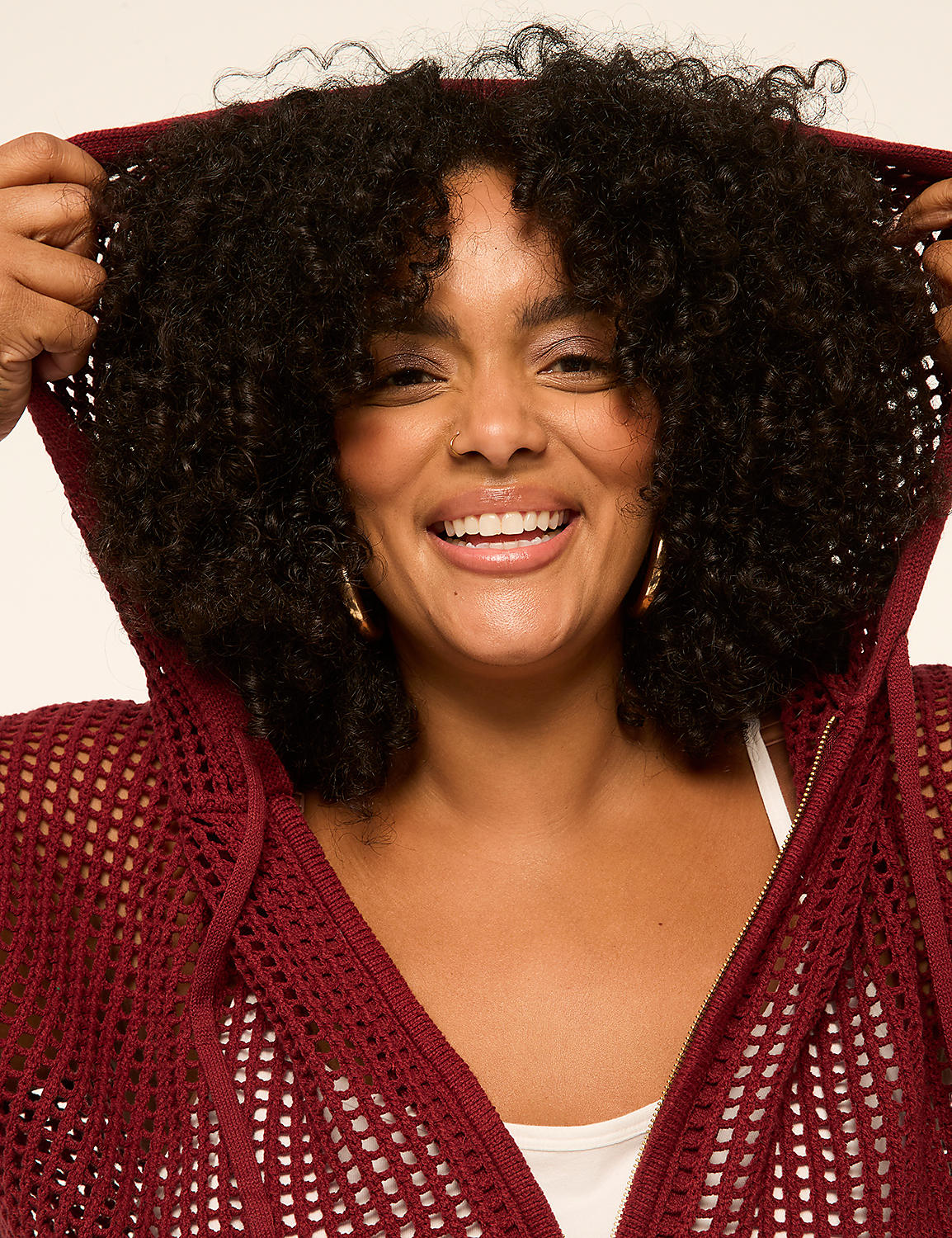 A smiling woman with curly hair pulls up the hood of her maroon crocheted cardigan against a plain background. Product Image 4