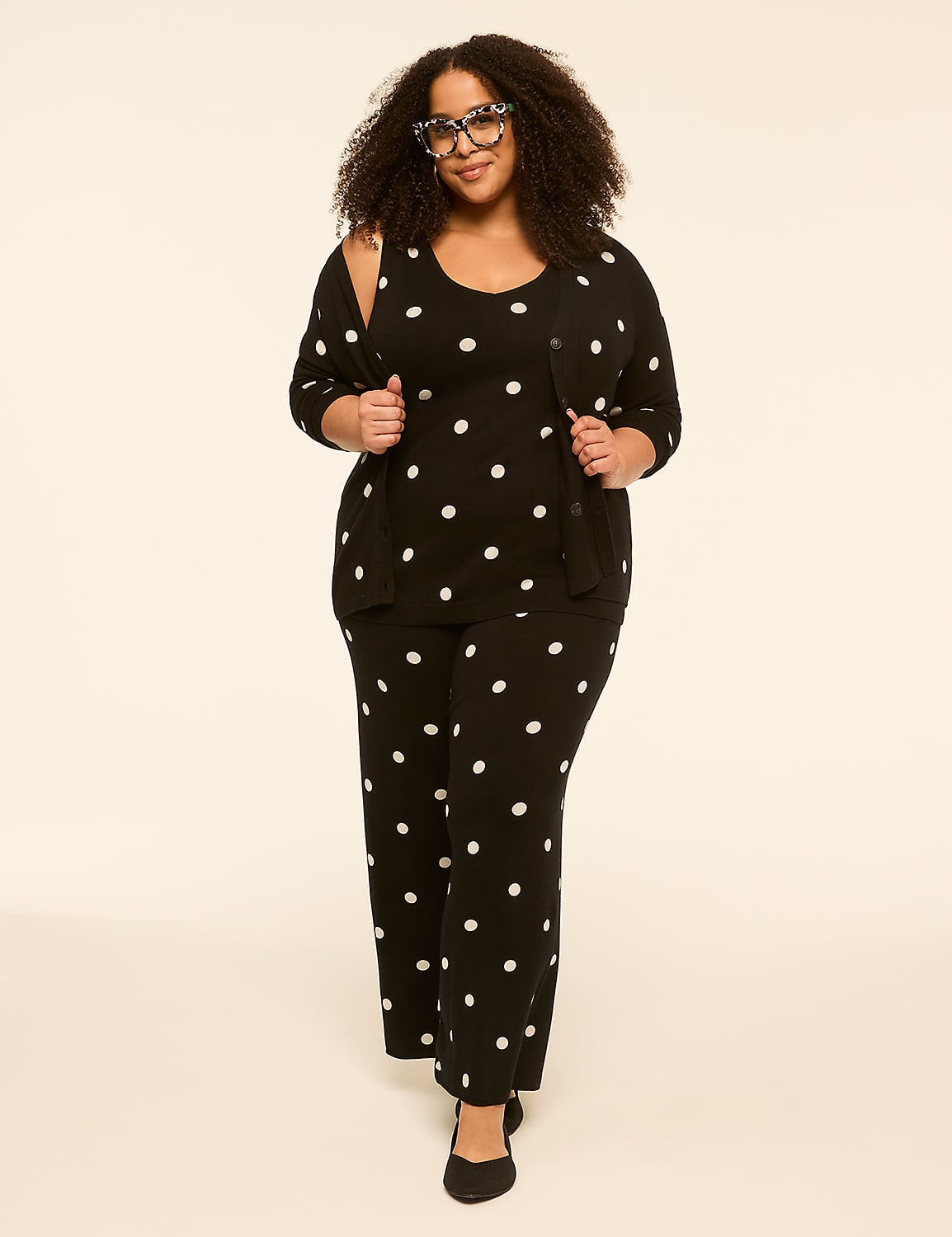 A woman with curly hair and glasses stands in the foreground wearing a black polka dot outfit, against a plain, light-colored background. Product Image 3
