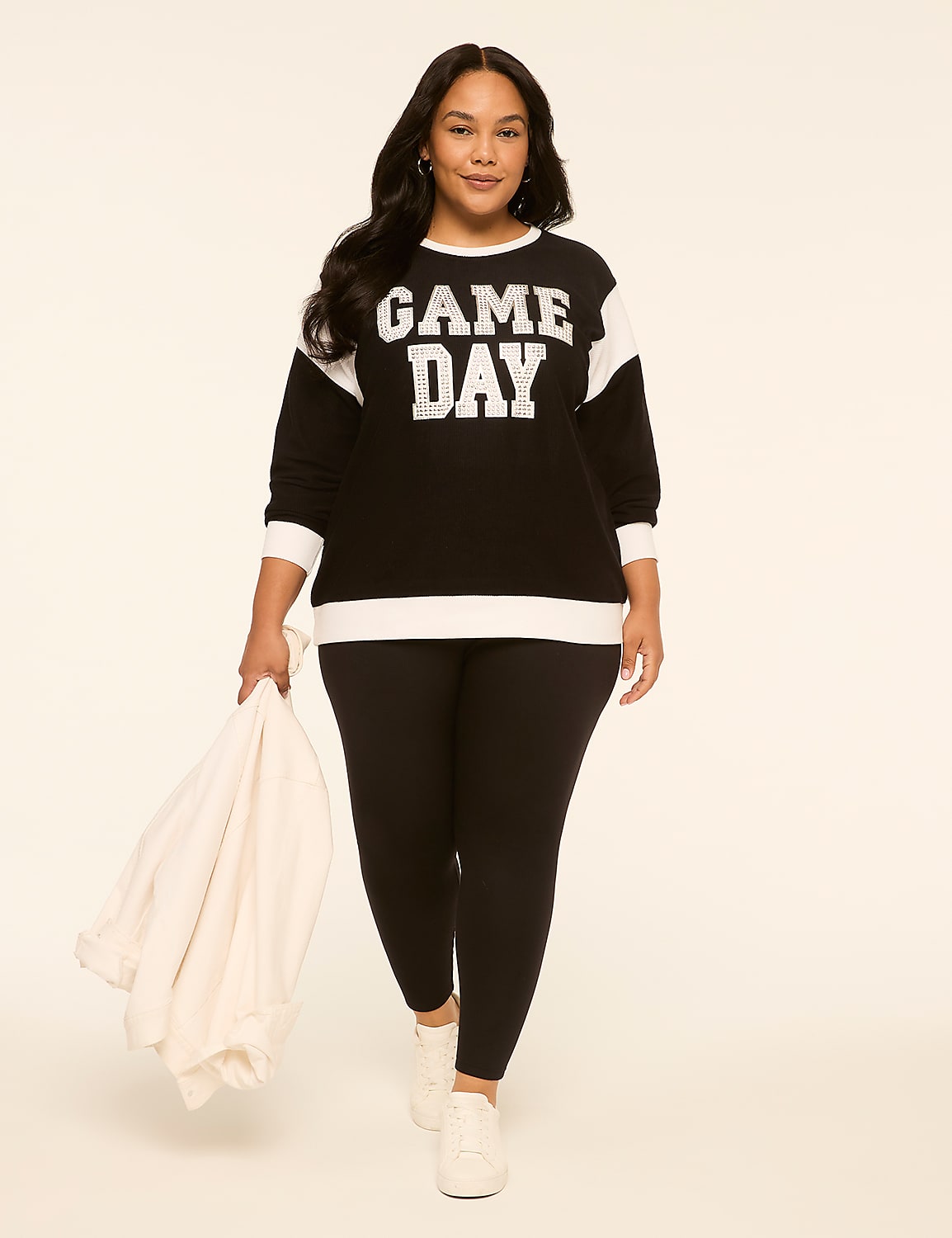 A woman in a black "GAME DAY" sweatshirt and black leggings walks forward, holding a white jacket, against a plain, light-colored background. Product Image 3