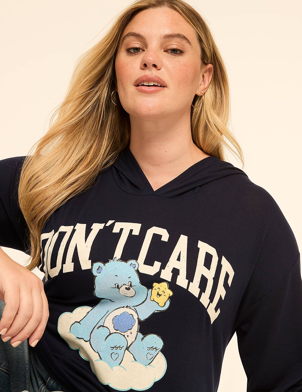 A woman with blonde hair wears a navy hoodie with a Care Bear graphic and the words "DON'T CARE" on it, against a plain, light-colored background. Product Image 4