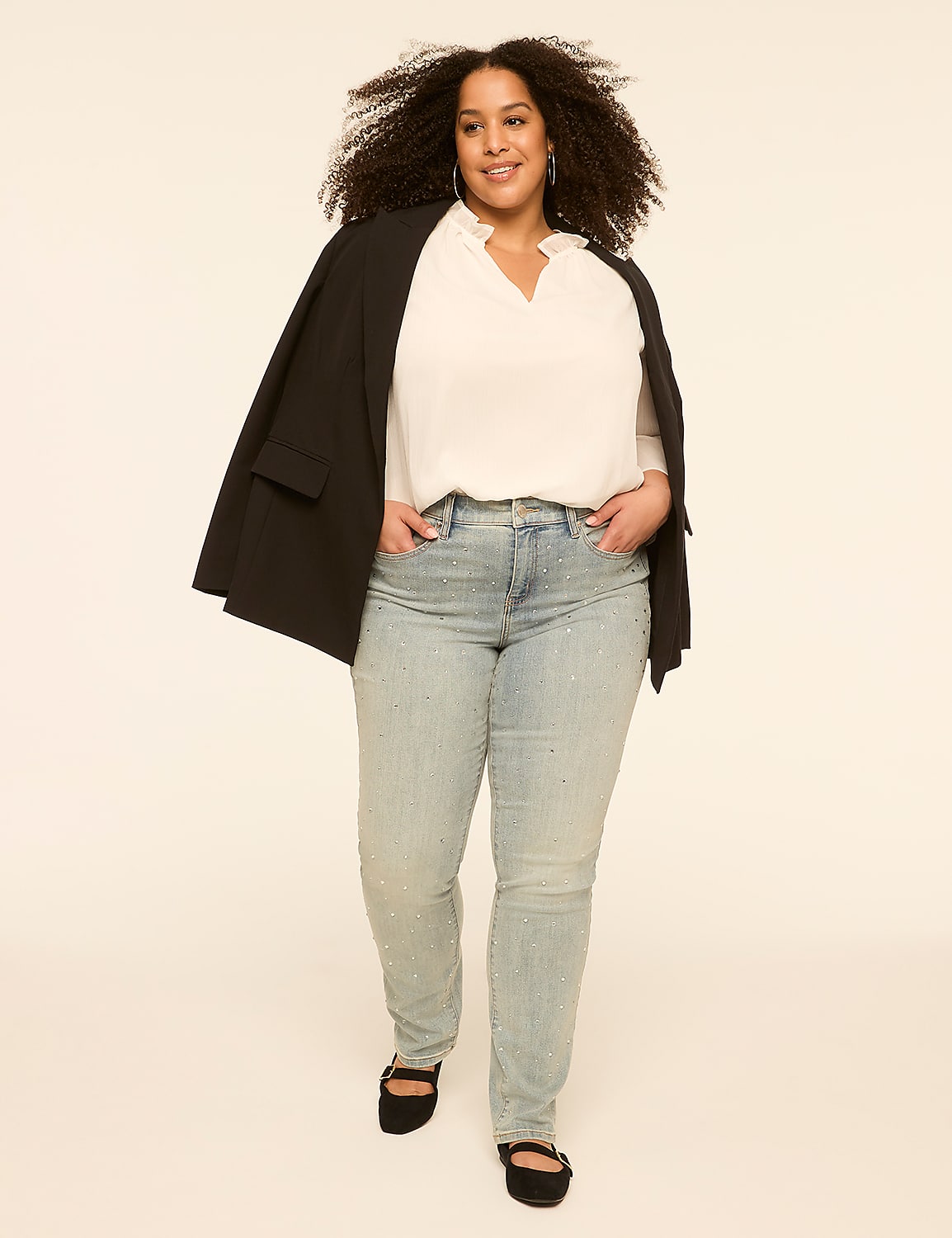 A woman with curly hair and a white blouse, light wash jeans, and a black blazer walks forward against a plain, light-colored background. Product Image 3