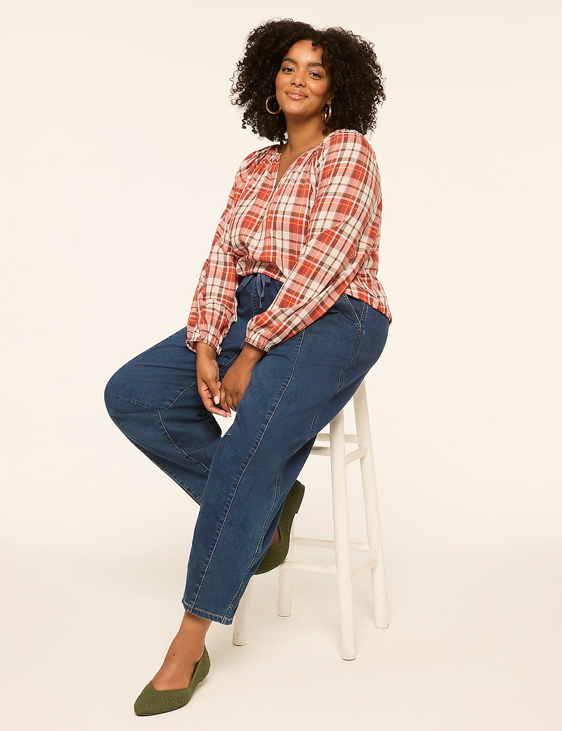 A woman with curly dark hair, wearing a plaid blouse and jeans, sits on a white stool against a plain cream background. Product Image 3