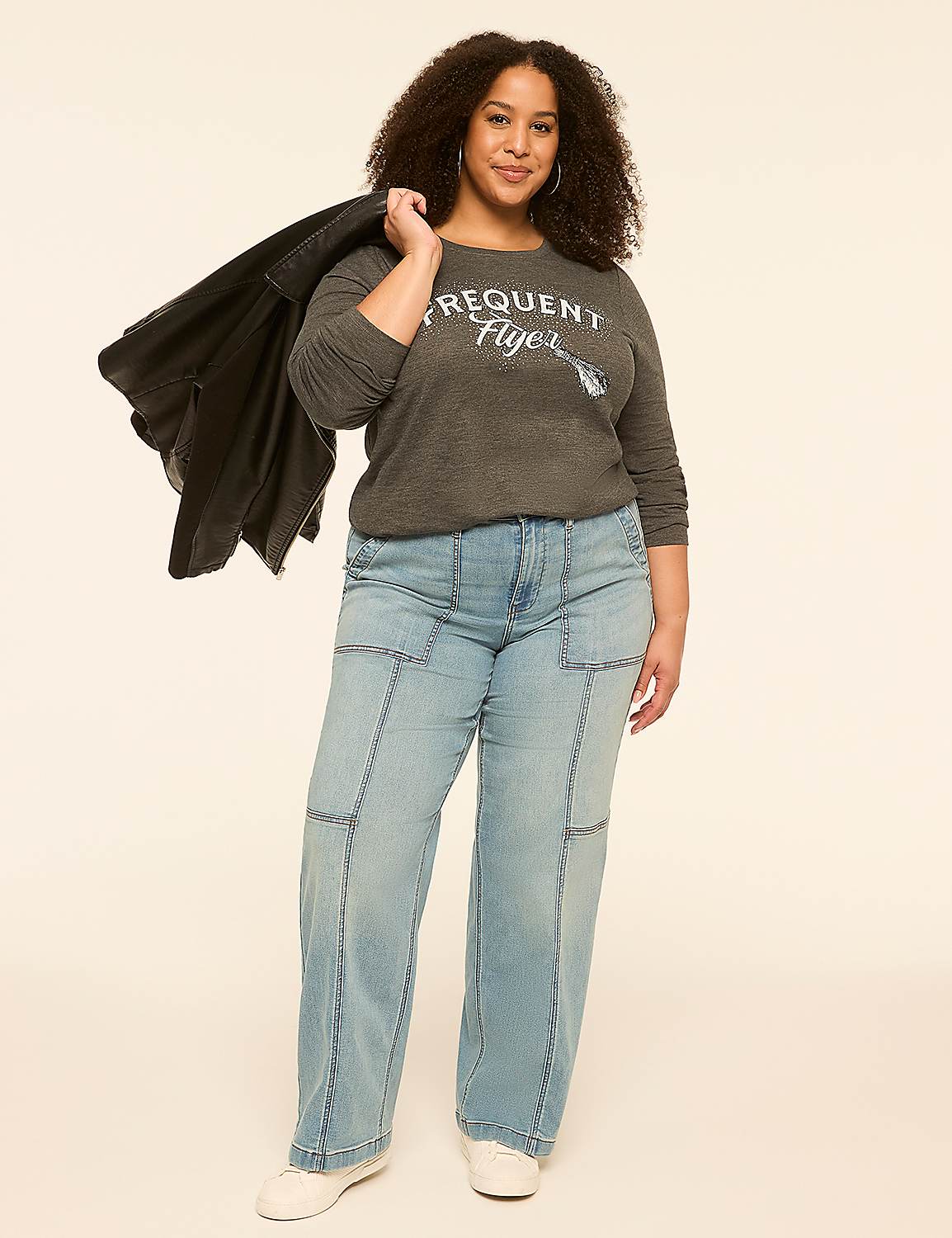 A woman in a gray sweatshirt and light blue jeans stands against a plain, light-colored background, with a black jacket draped over her shoulder. Product Image 3