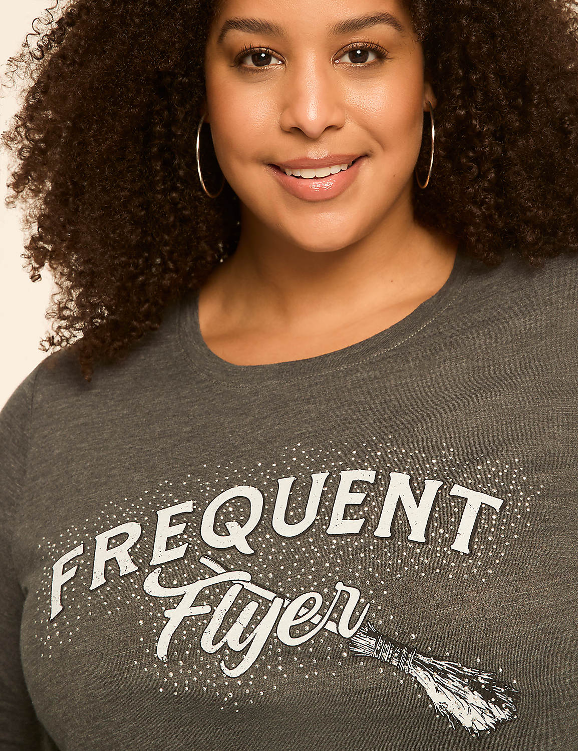 A smiling woman with curly hair wears a grey t-shirt with "Frequent Flyer" and a broomstick graphic, against a plain, light-colored background. Product Image 4