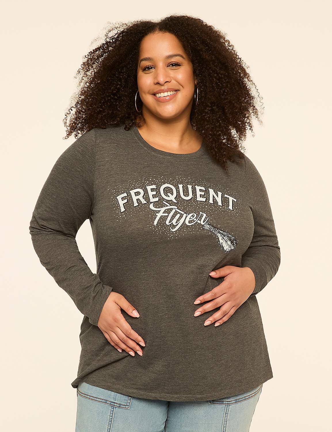 A smiling woman with curly hair in a grey long-sleeved shirt with "Frequent Flyer" printed on it stands against a plain, light-colored background. Product Image 1