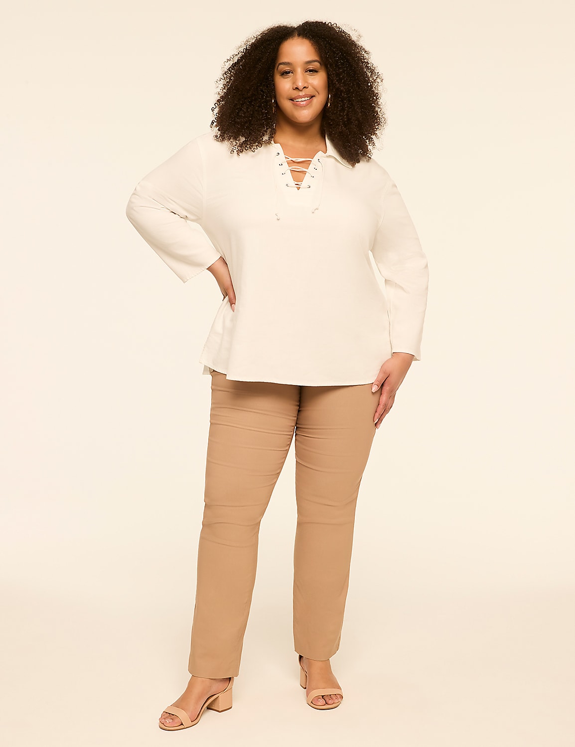 A smiling woman with curly dark hair stands in a plain, light-colored studio, wearing a white lace-up top and tan pants. Product Image 3