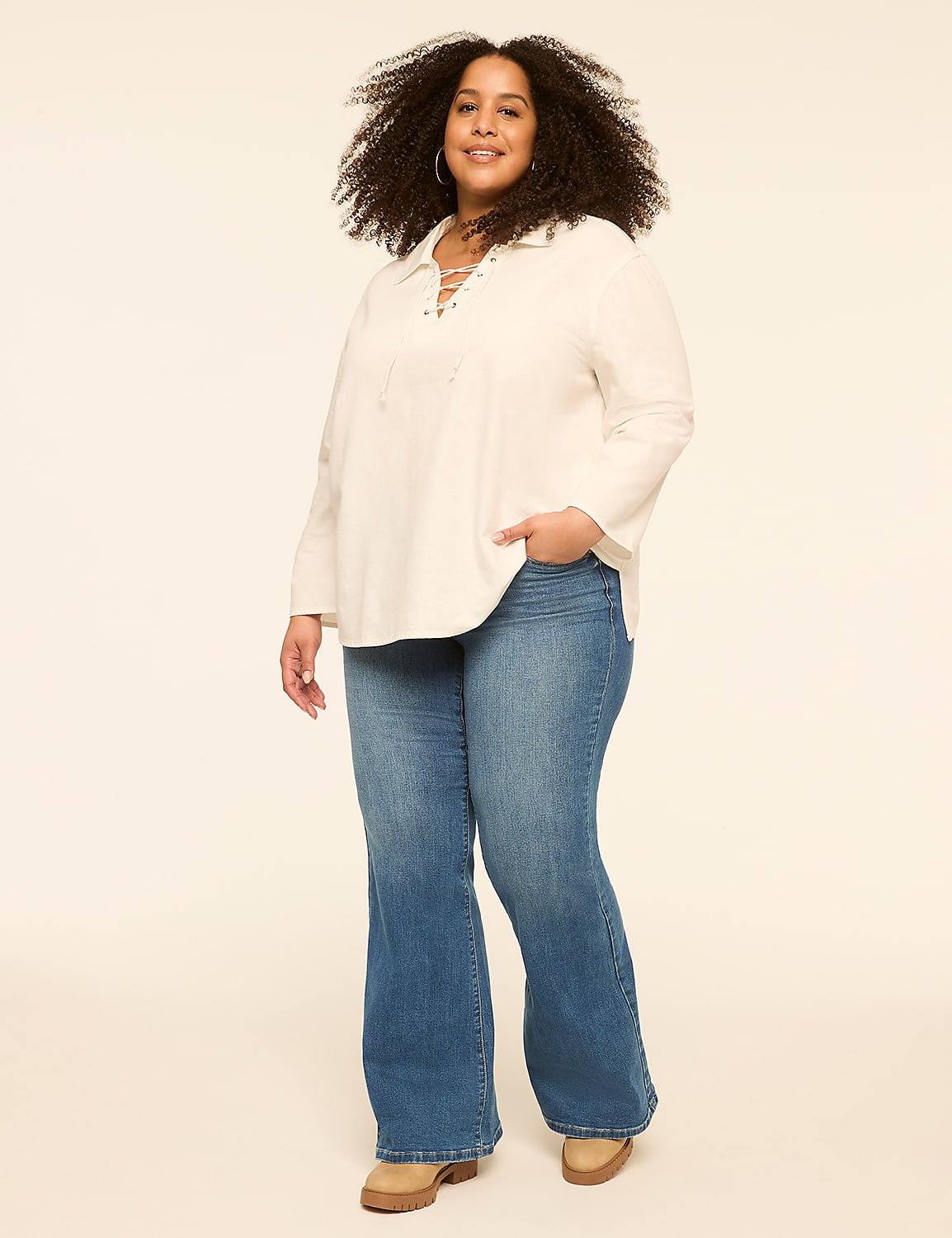 A woman with curly hair stands in the foreground wearing a white lace-up top and flared jeans against a plain, light-colored background. Product Image 4