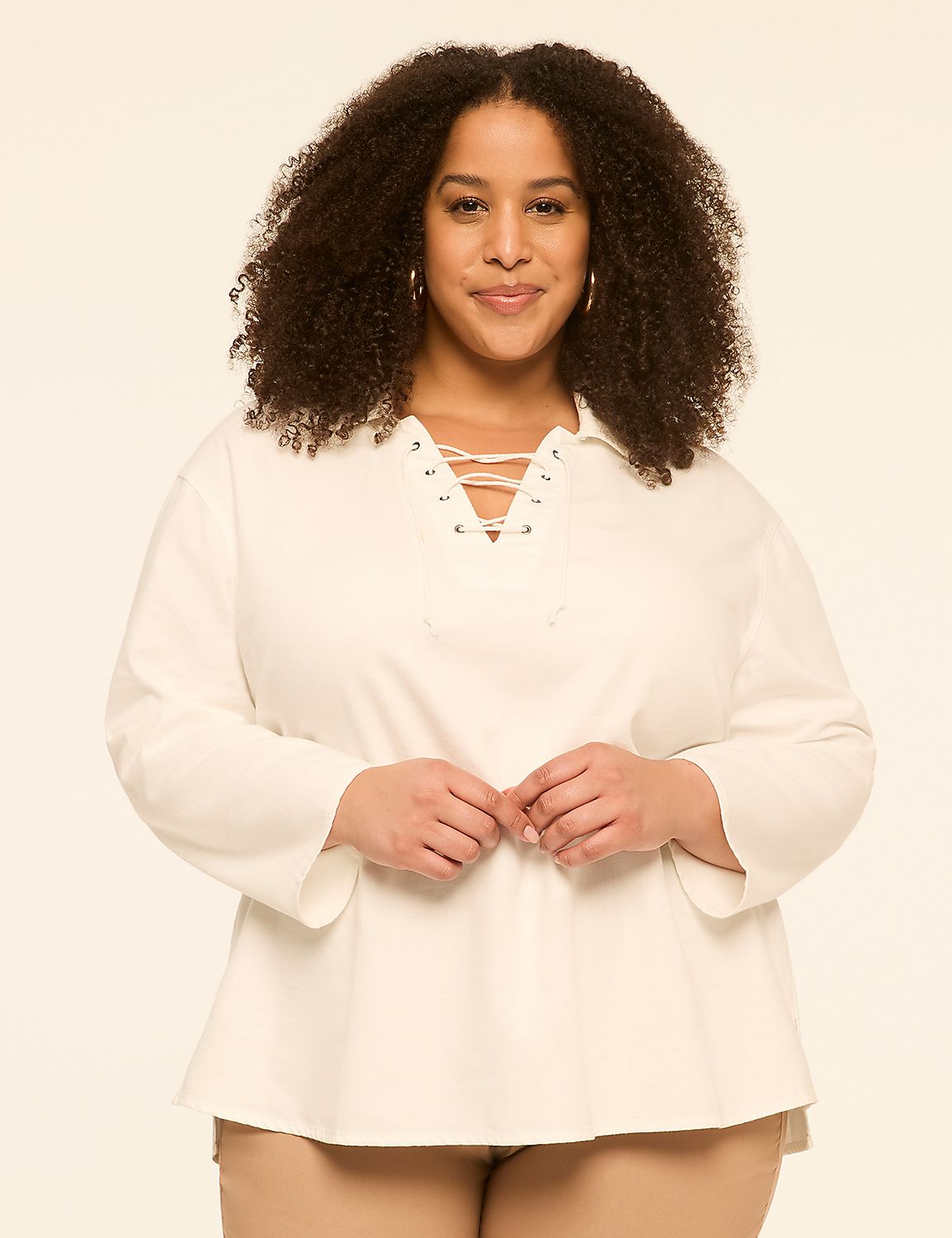 A woman with curly dark hair and a white lace-up shirt stands against a plain, light-colored background. Product Image 1