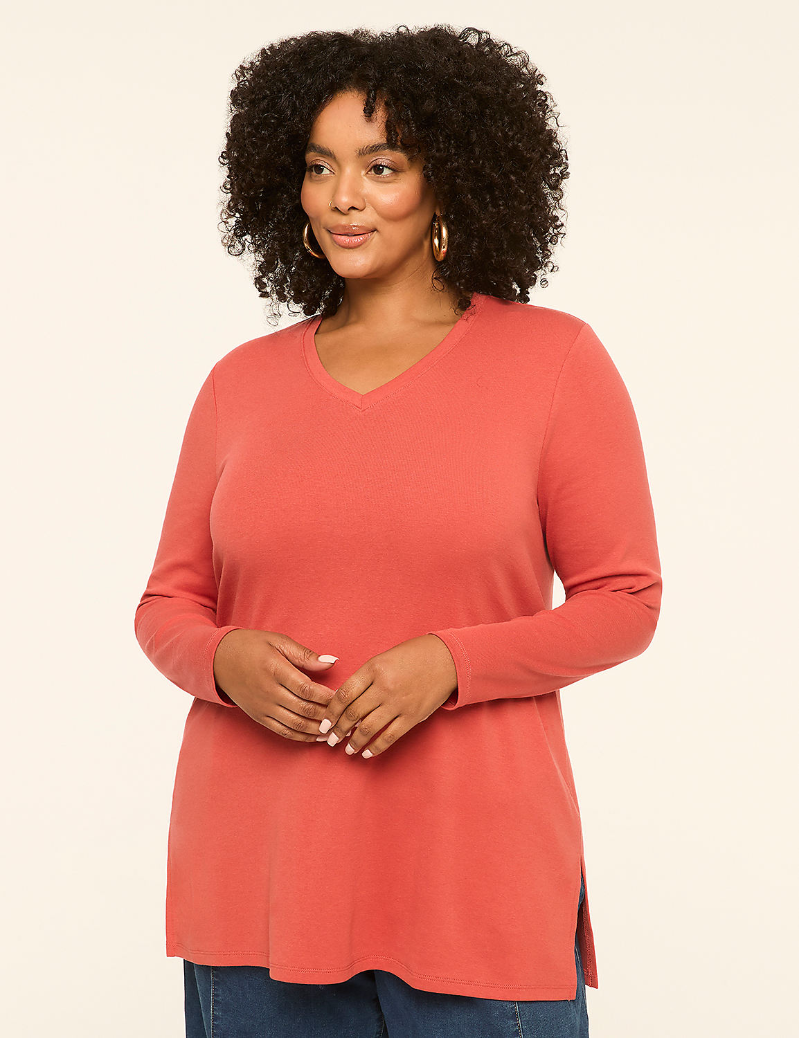 A woman with curly dark hair and hoop earrings poses in a coral v-neck shirt against a plain cream background. Product Image 1