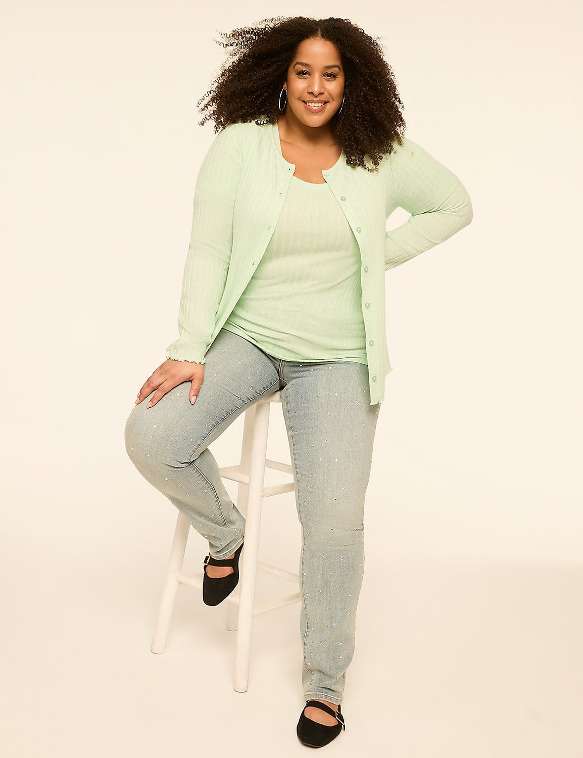 A woman with curly hair sits on a white stool in the foreground, wearing a light green cardigan and jeans, against a plain, light-colored background. Product Image 3