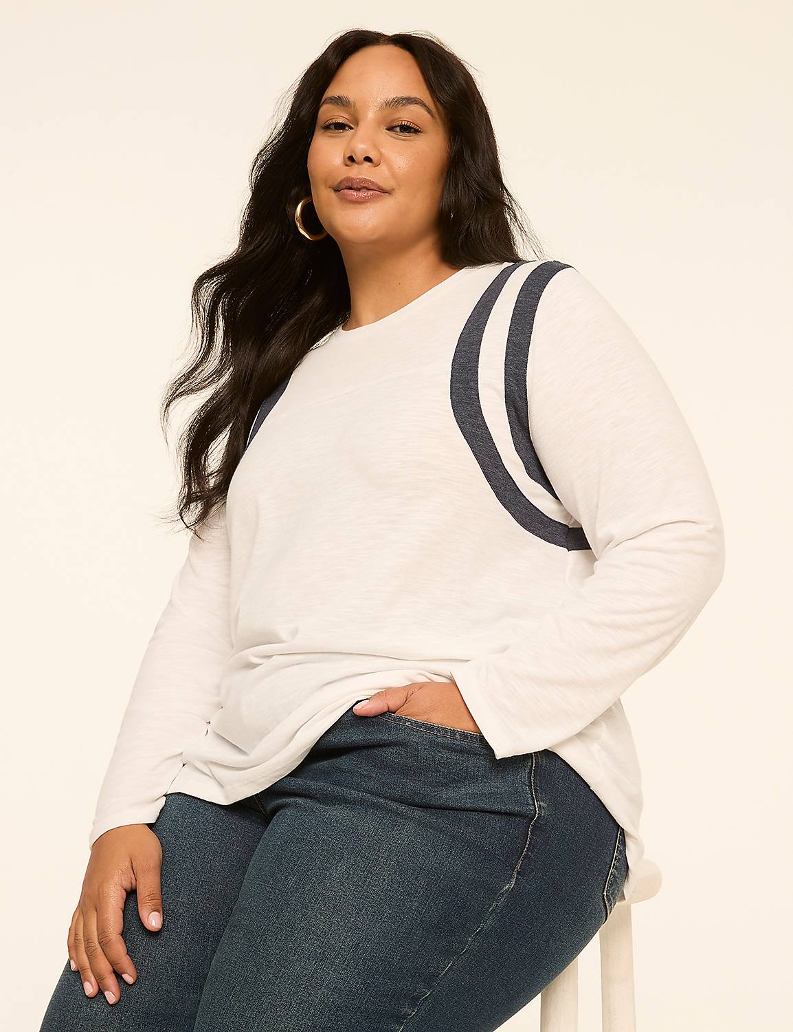 A woman with dark hair and hoop earrings poses in a white long-sleeved shirt with navy stripes on the shoulders and dark wash jeans against a plain, light-colored background. Product Image 1