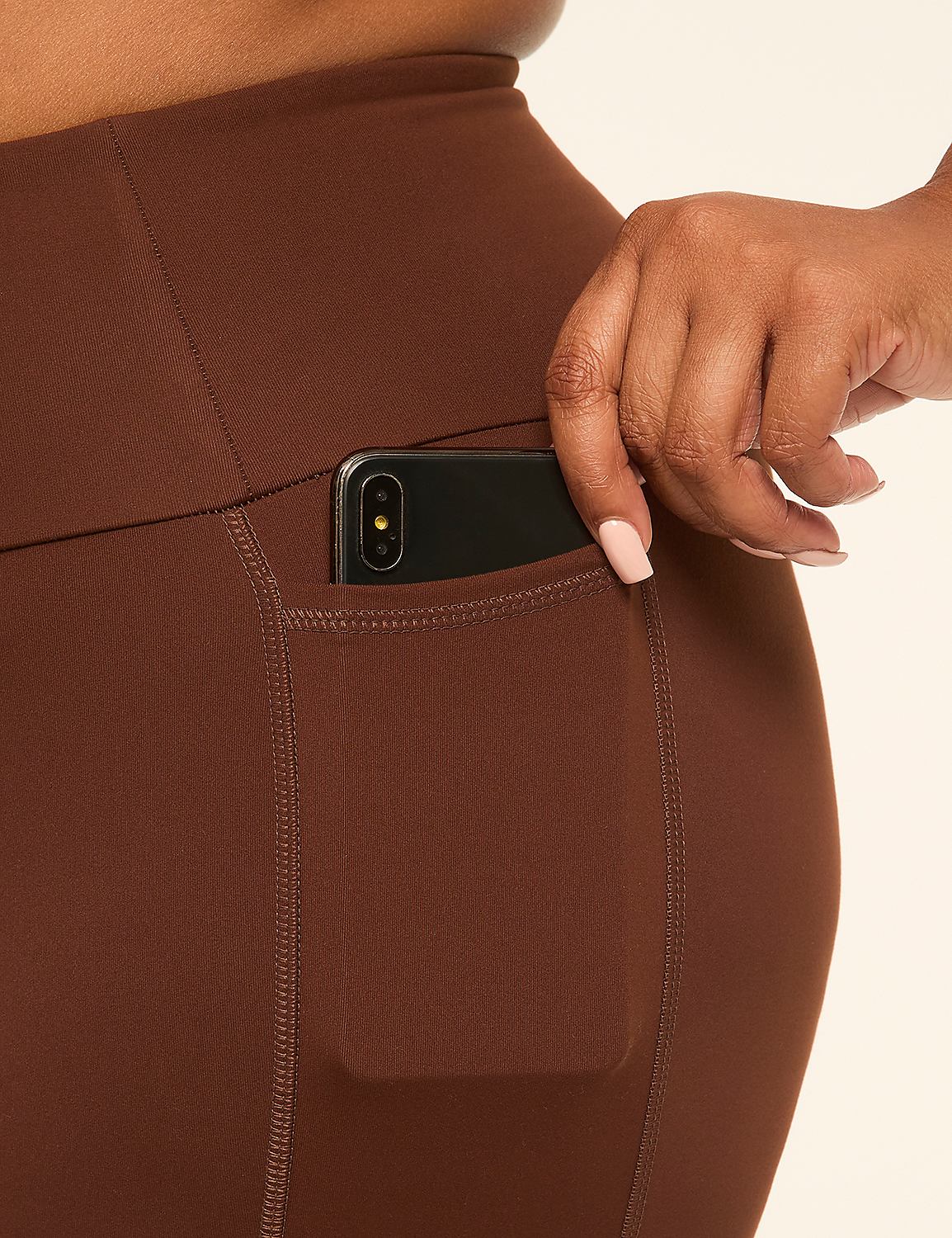 A hand with a light pink manicure places a smartphone into the pocket of brown leggings against a plain cream background. Product Image 4