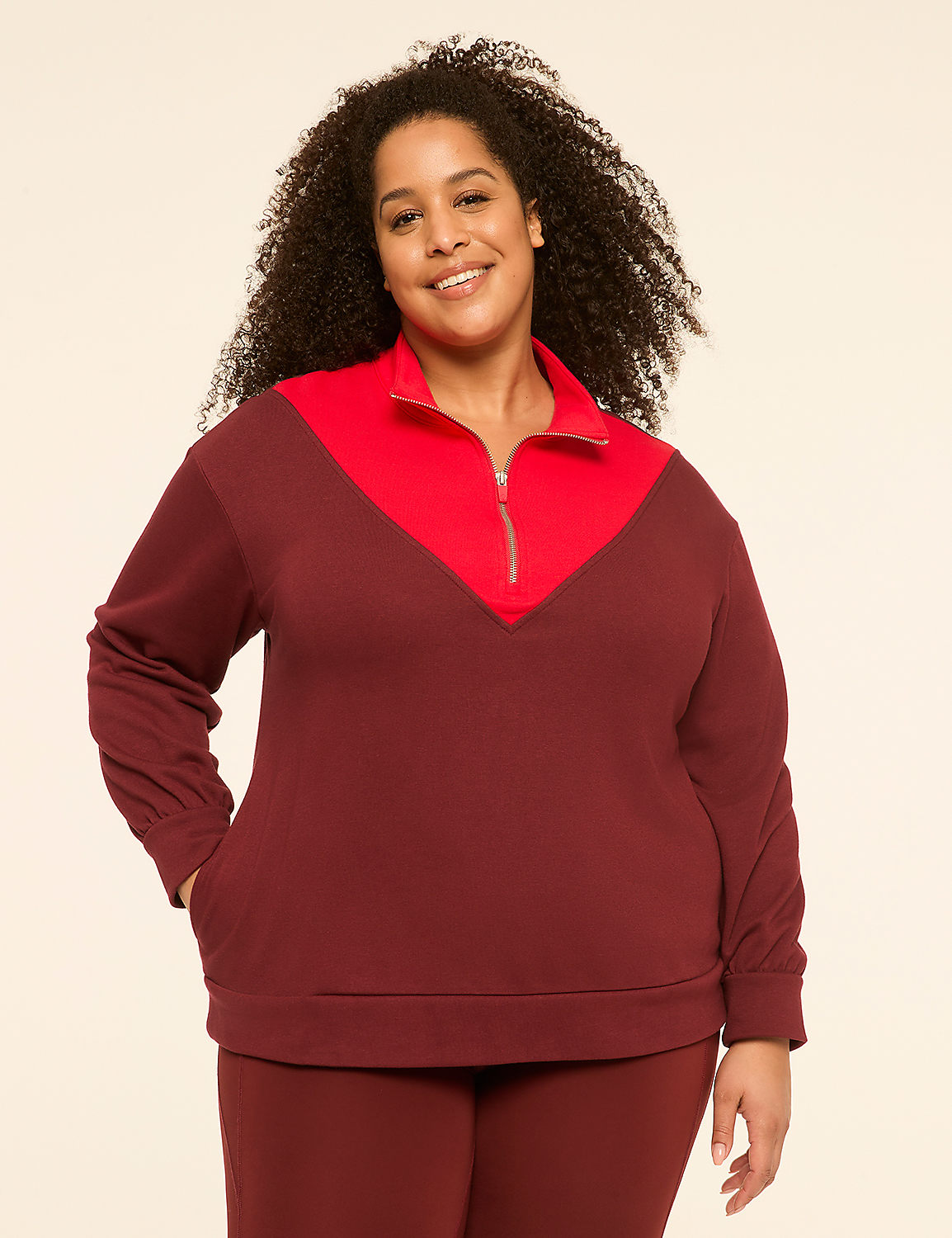 A smiling woman with curly hair stands in the foreground wearing a maroon and red quarter-zip sweatshirt and matching pants, against a plain cream-colored background. Product Image 1
