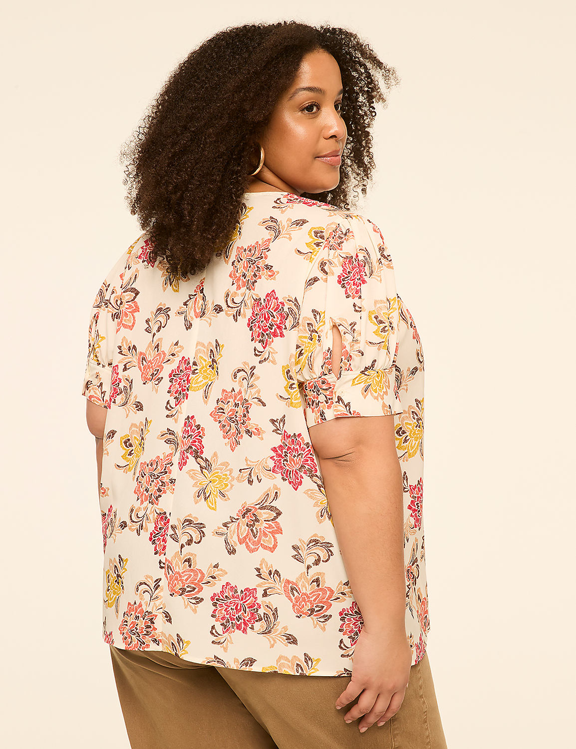 A woman with curly hair wears a floral blouse and brown pants against a plain background. Product Image 2