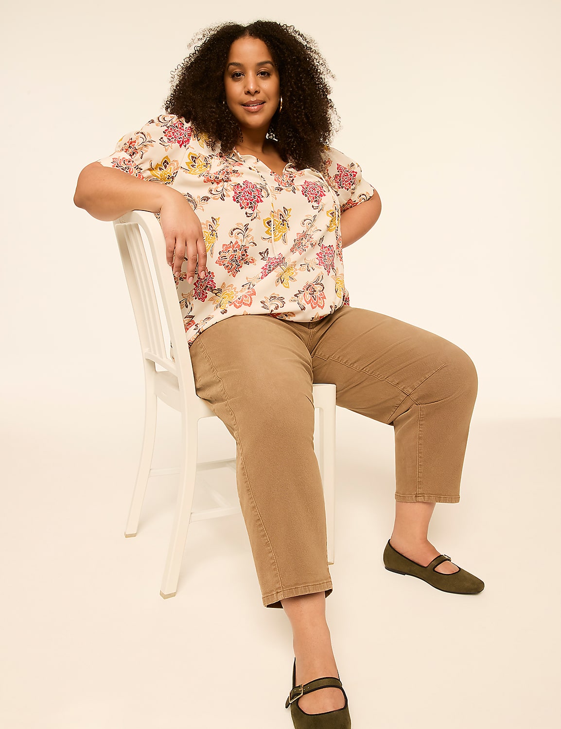 A woman with curly hair sits on a white chair in the foreground, wearing a floral blouse and brown pants, against a plain, light-colored background. Product Image 3