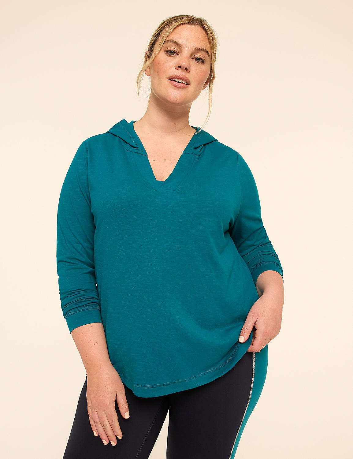 A woman in a teal hooded shirt and dark leggings stands against a plain beige background. Product Image 1