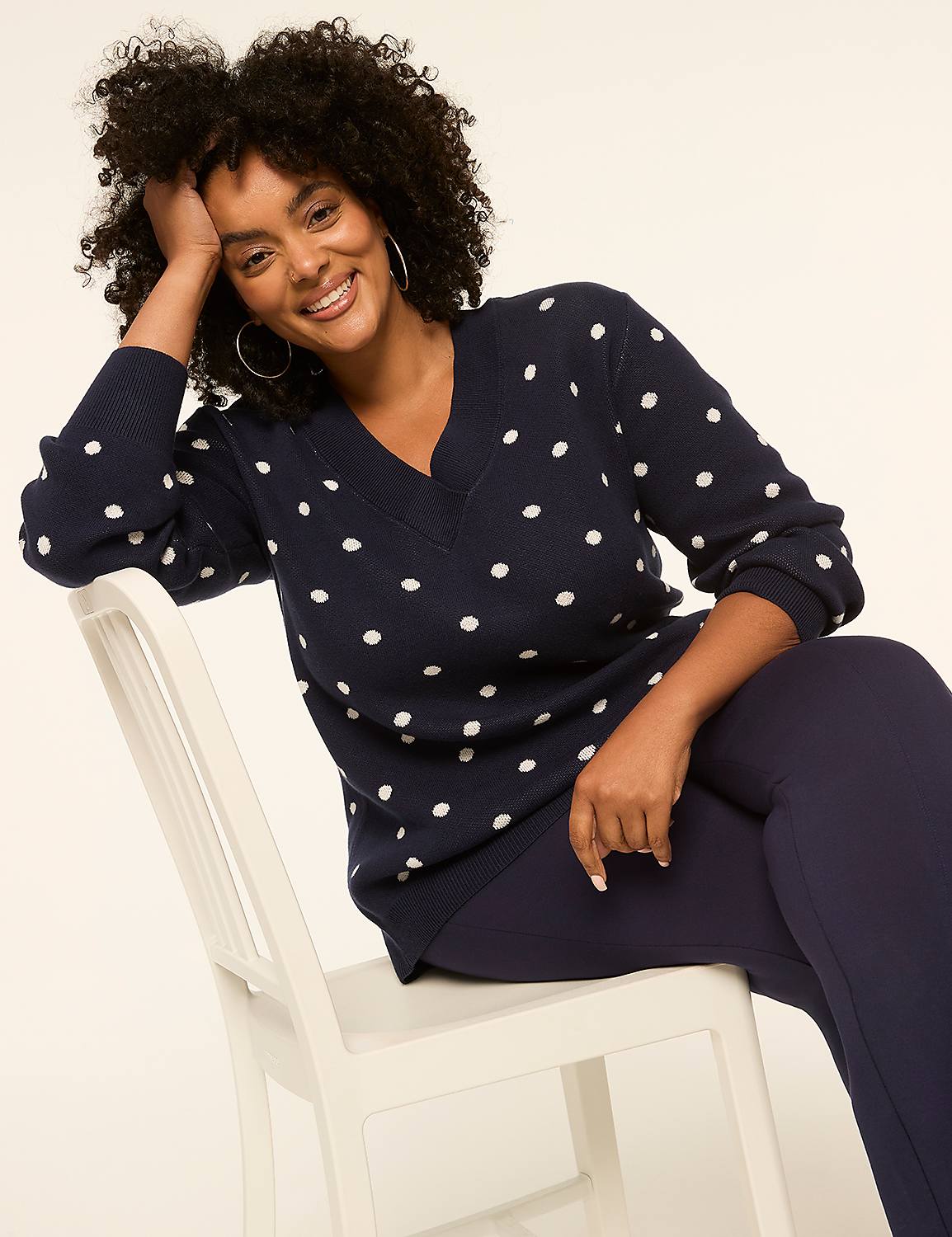 A smiling woman with dark curly hair, wearing a navy polka dot sweater and dark leggings, sits on a white chair against a plain, light background. Product Image 3
