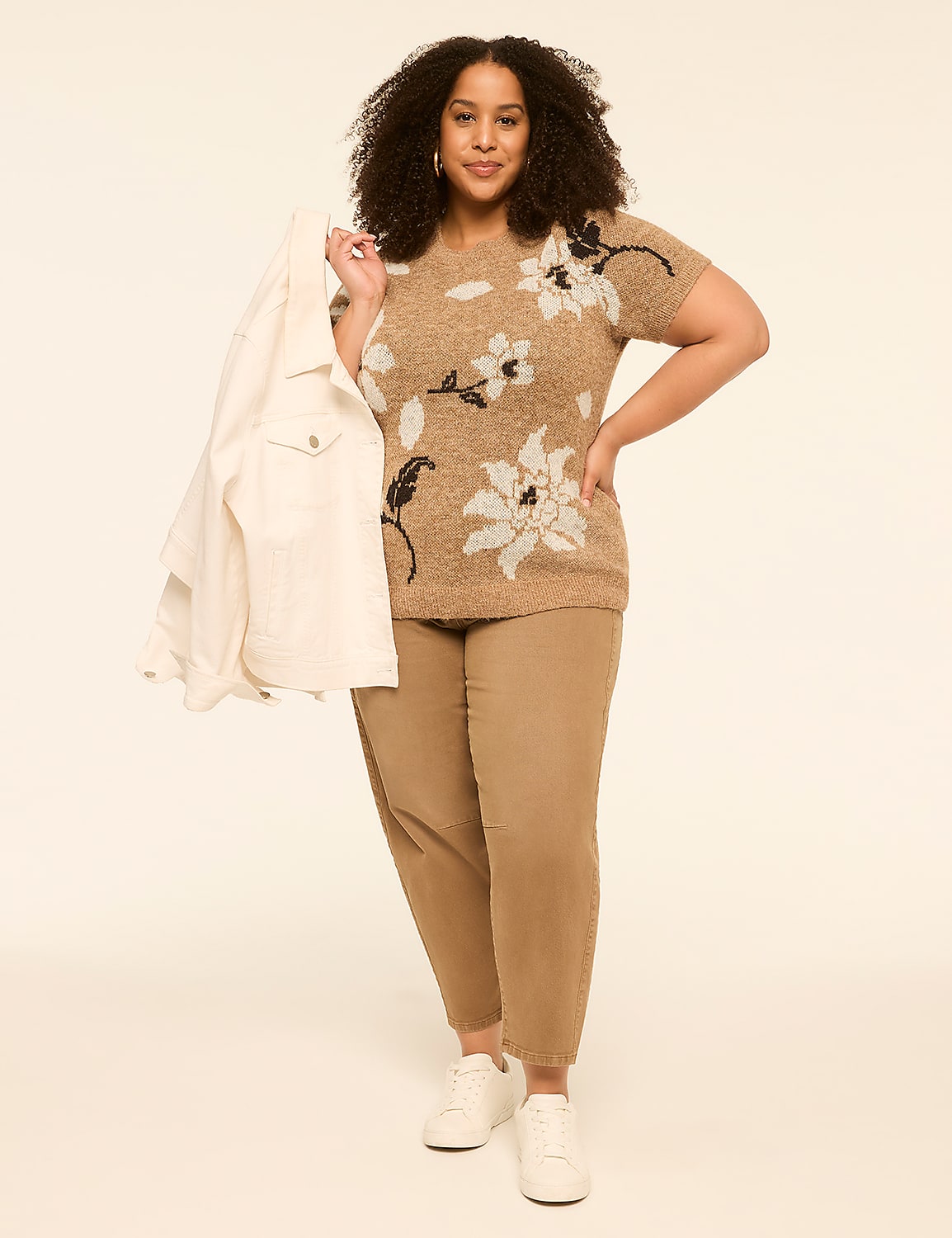 A woman in a floral sweater and tan pants stands against a plain, light background, holding a white jacket. Product Image 3