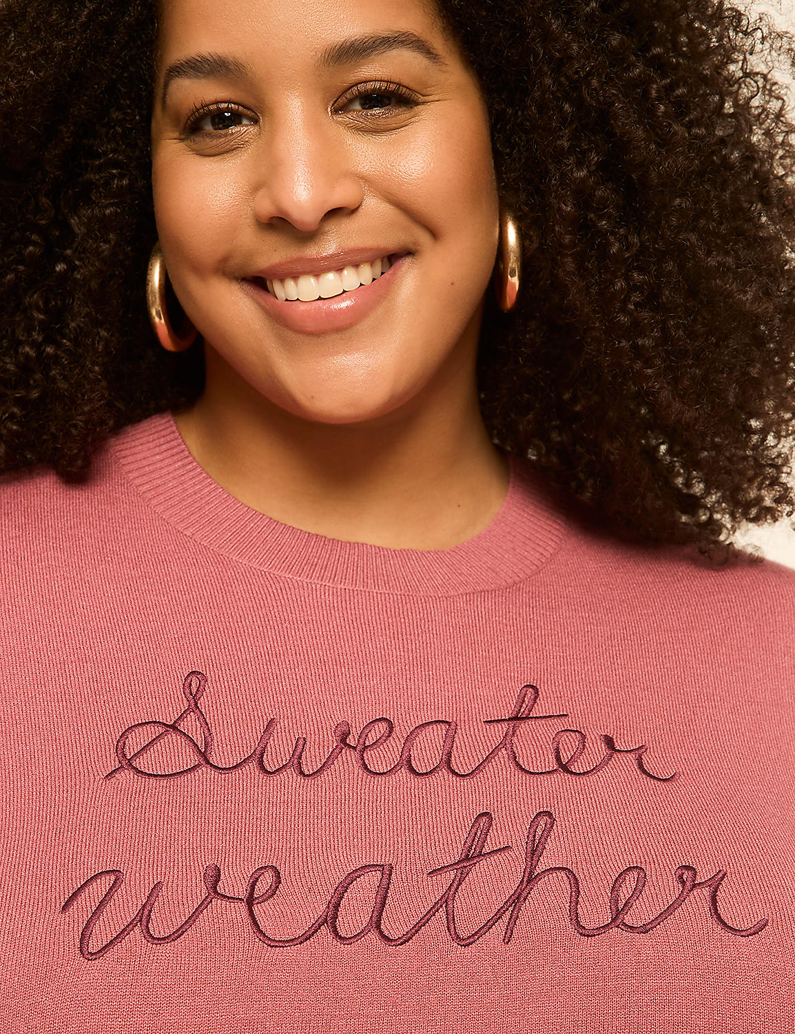 A smiling woman with curly hair wears a pink sweater with "Sweater weather" embroidered on it, against a plain background. Product Image 4