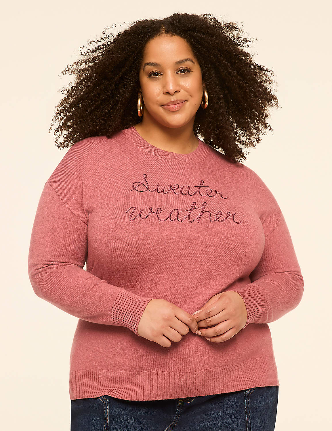 A woman with curly hair smiles in a pink "Sweater weather" sweatshirt against a plain, light-colored background. Product Image 1