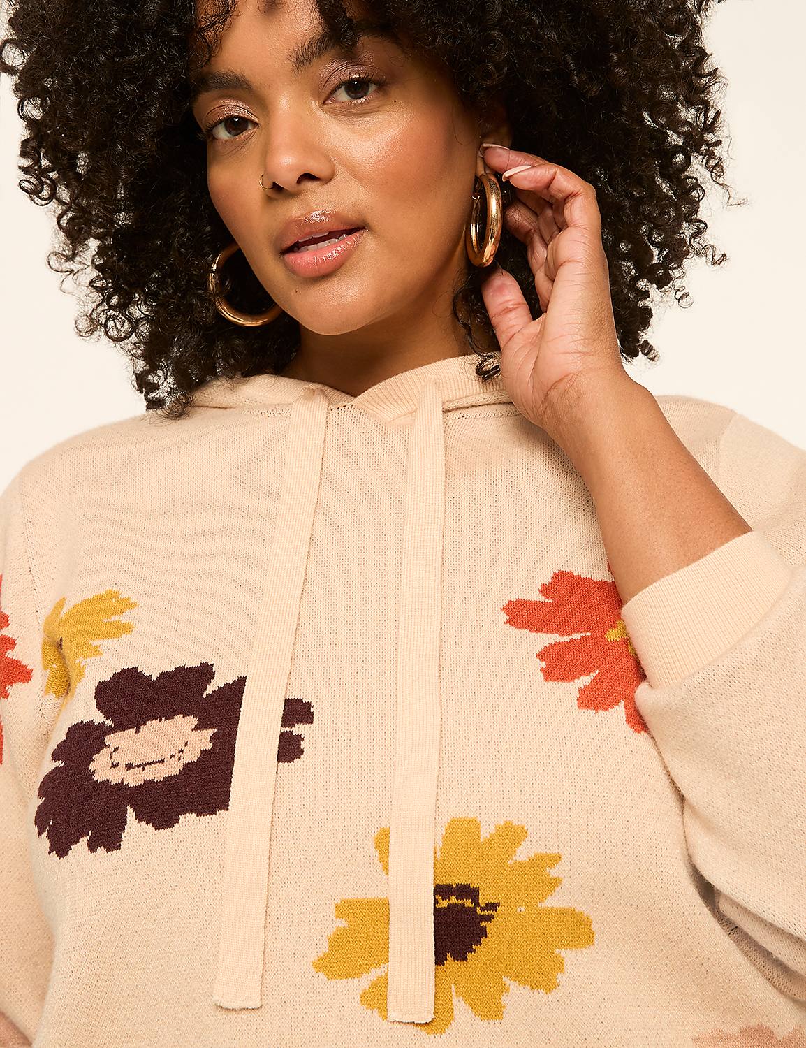 A woman with curly hair and large hoop earrings poses in a cream-colored hoodie with a floral pattern against a plain background. Product Image 4