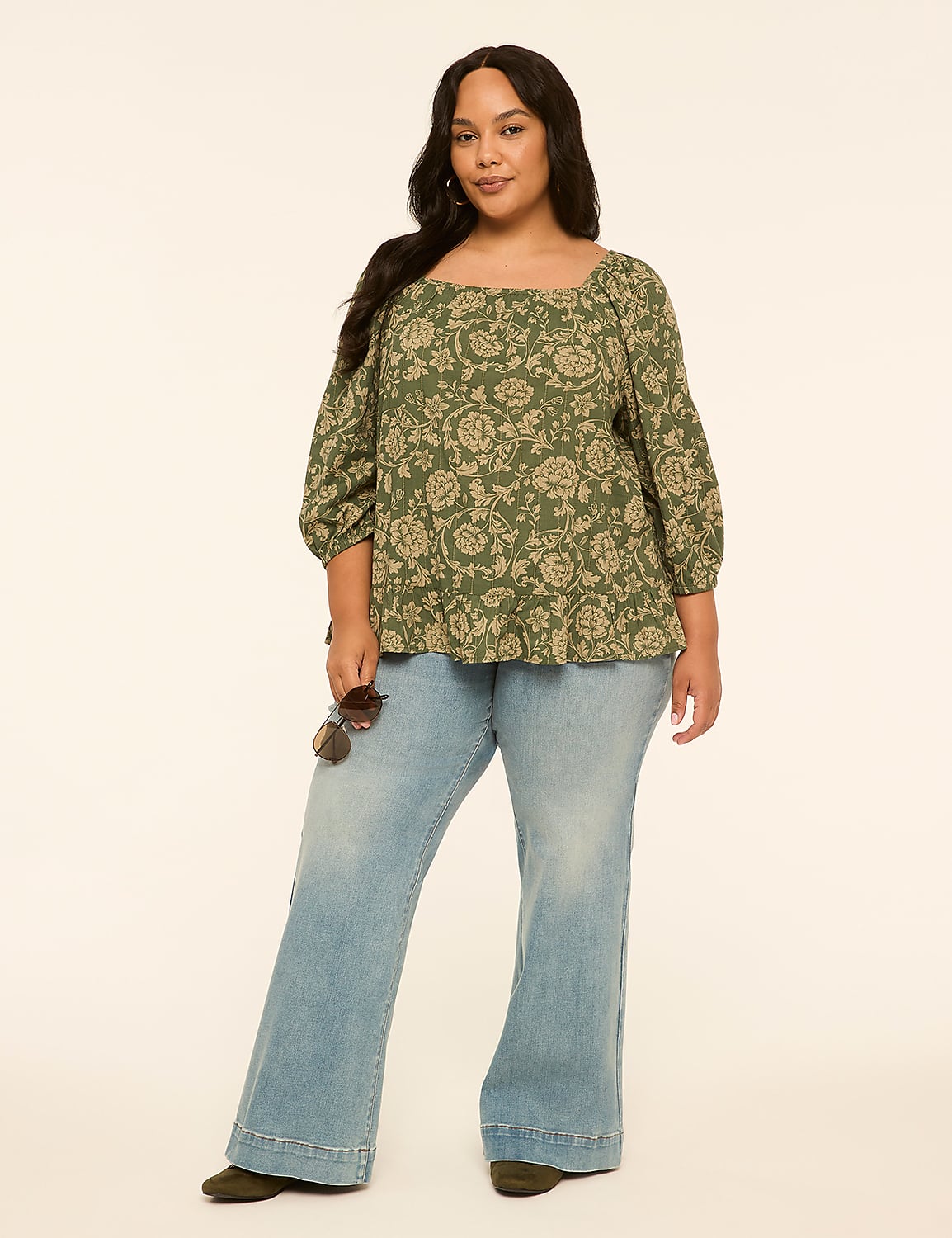 A woman in a floral top and flared jeans stands against a plain, light-colored background, holding sunglasses. Product Image 3