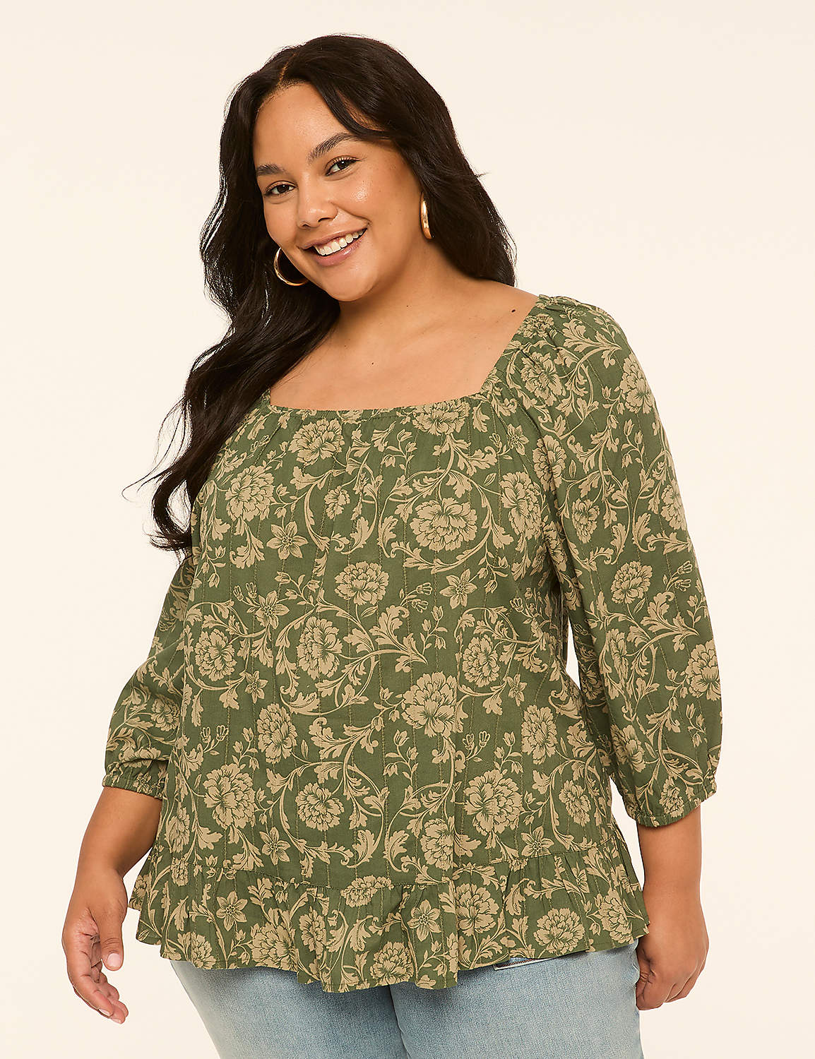 A smiling woman with dark hair and hoop earrings wears a green floral-print peasant blouse with a square neckline and light blue jeans, set against a plain cream background. Product Image 1