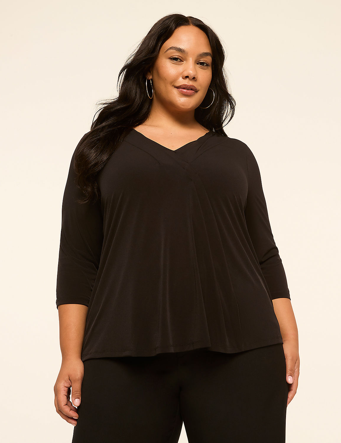A woman with dark hair and hoop earrings poses in a black v-neck top and black pants against a plain beige background. Product Image 1