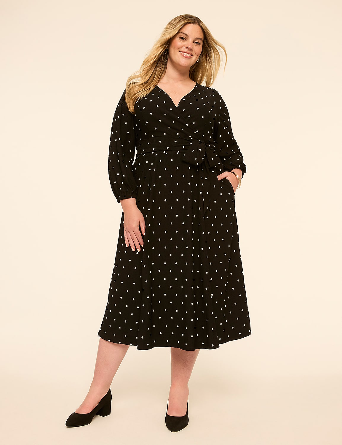 A smiling woman in a black polka-dot wrap dress stands against a plain, light-colored background. Product Image 1