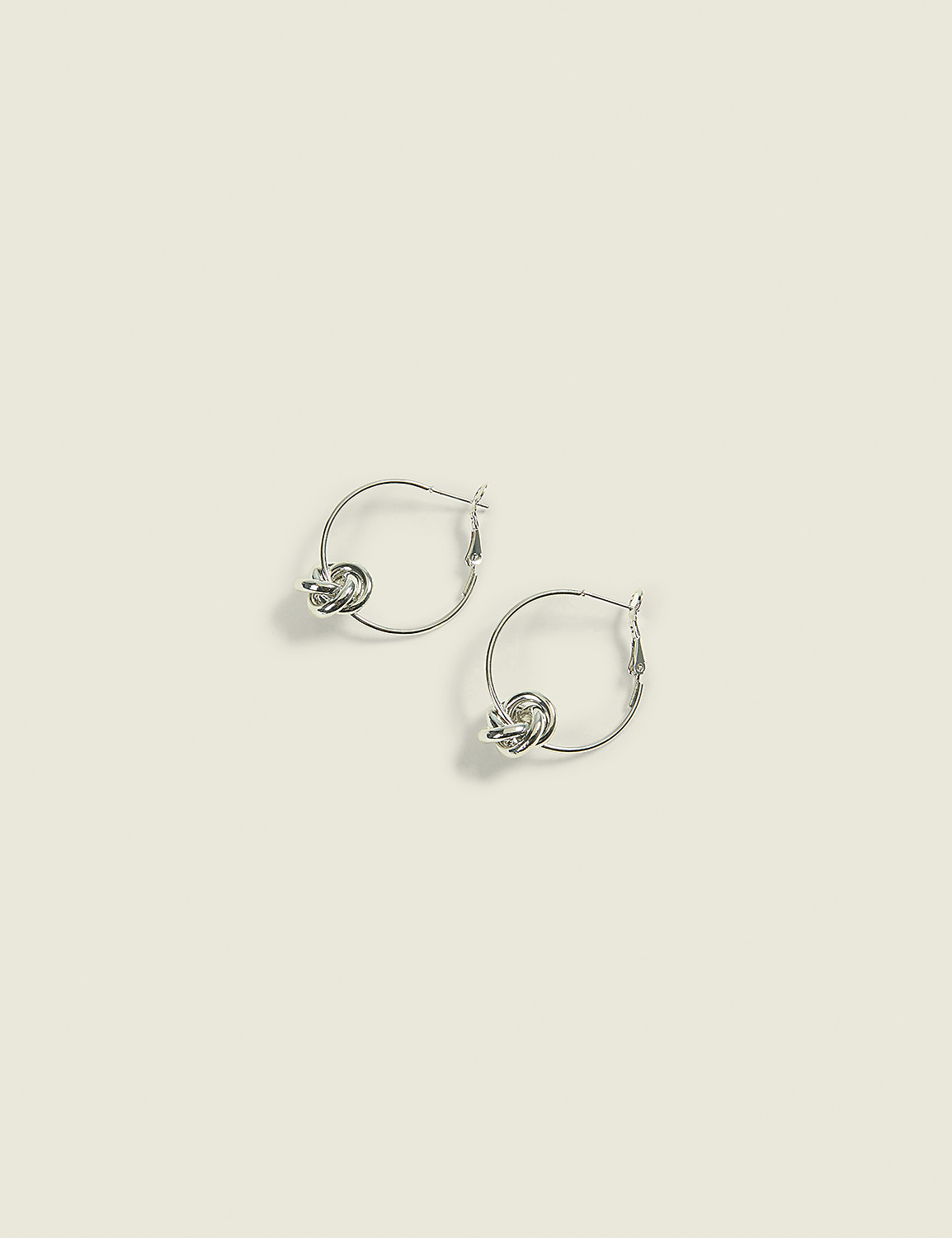 A pair of silver hoop earrings with knot details rest on a pale, textured background. Product Image 1
