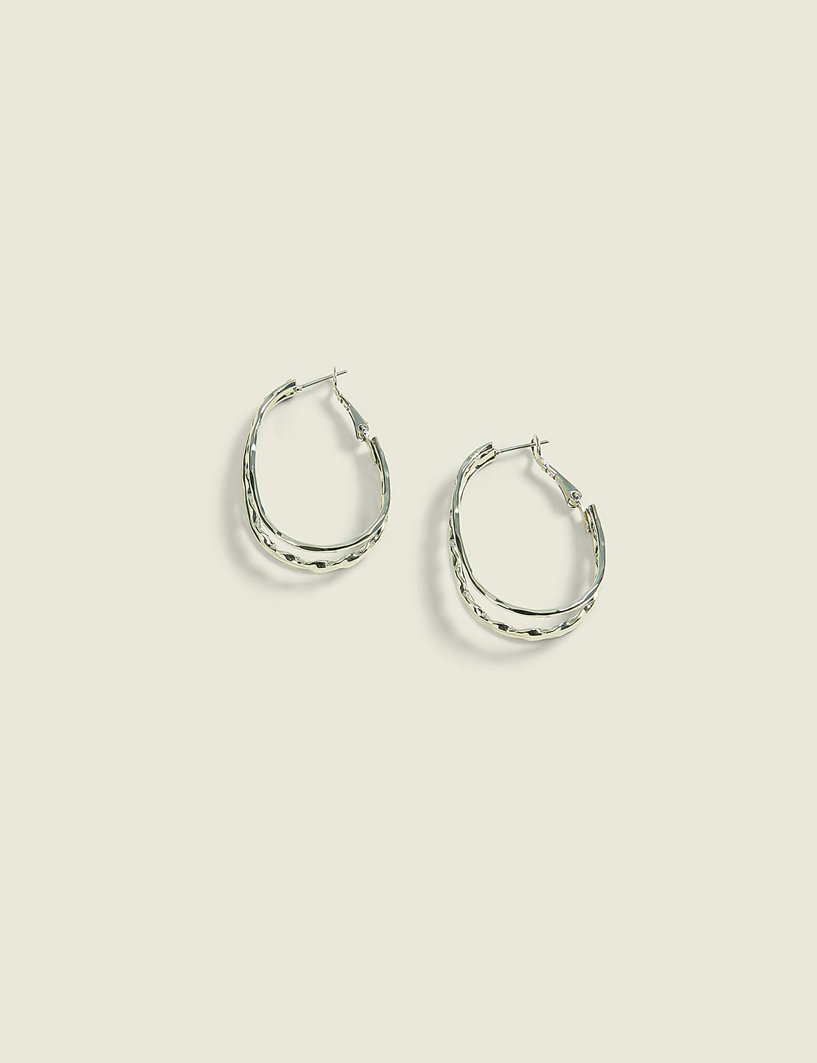 A pair of silver oval hoop earrings with textured detailing rests on a pale green background. Product Image 1