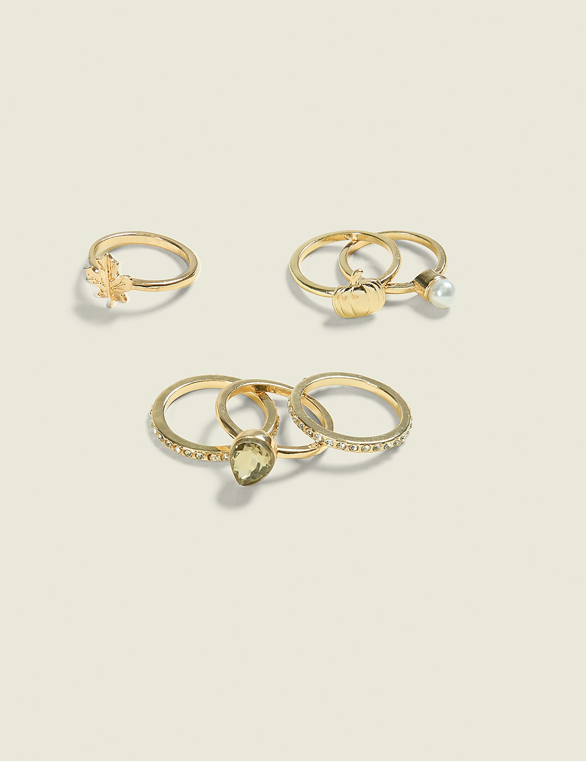 A collection of gold rings, including one with a leaf design, a pumpkin and pearl ring, and a set with a green stone and rhinestones, are arranged on a pale, textured background. Product Image 1