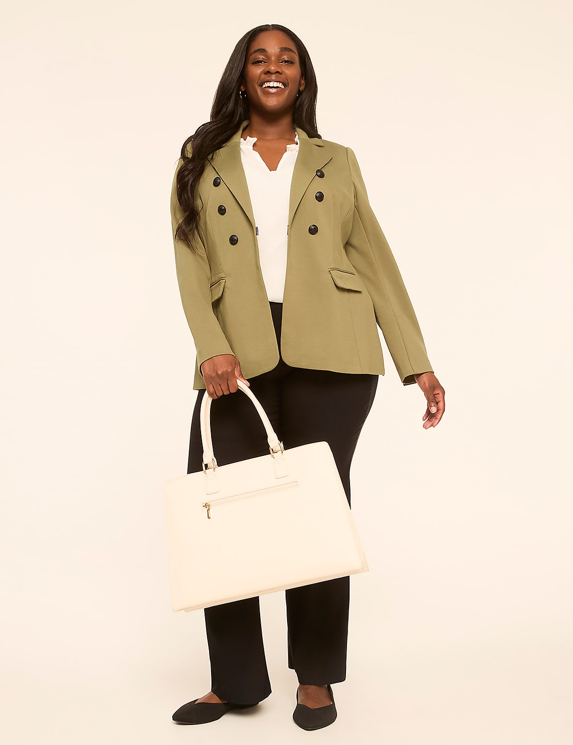 A smiling woman in a green blazer and black pants holds a cream-colored tote bag against a plain, light background. Product Image 3