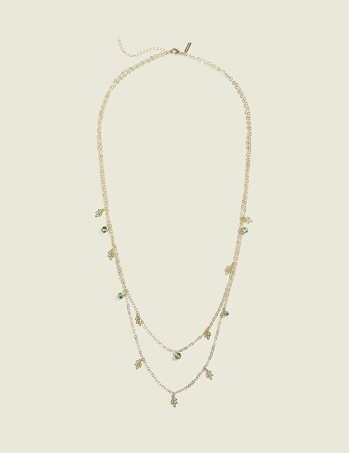 A delicate gold layered necklace with small green gemstone charms hangs against a plain, light beige background. Product Image 1