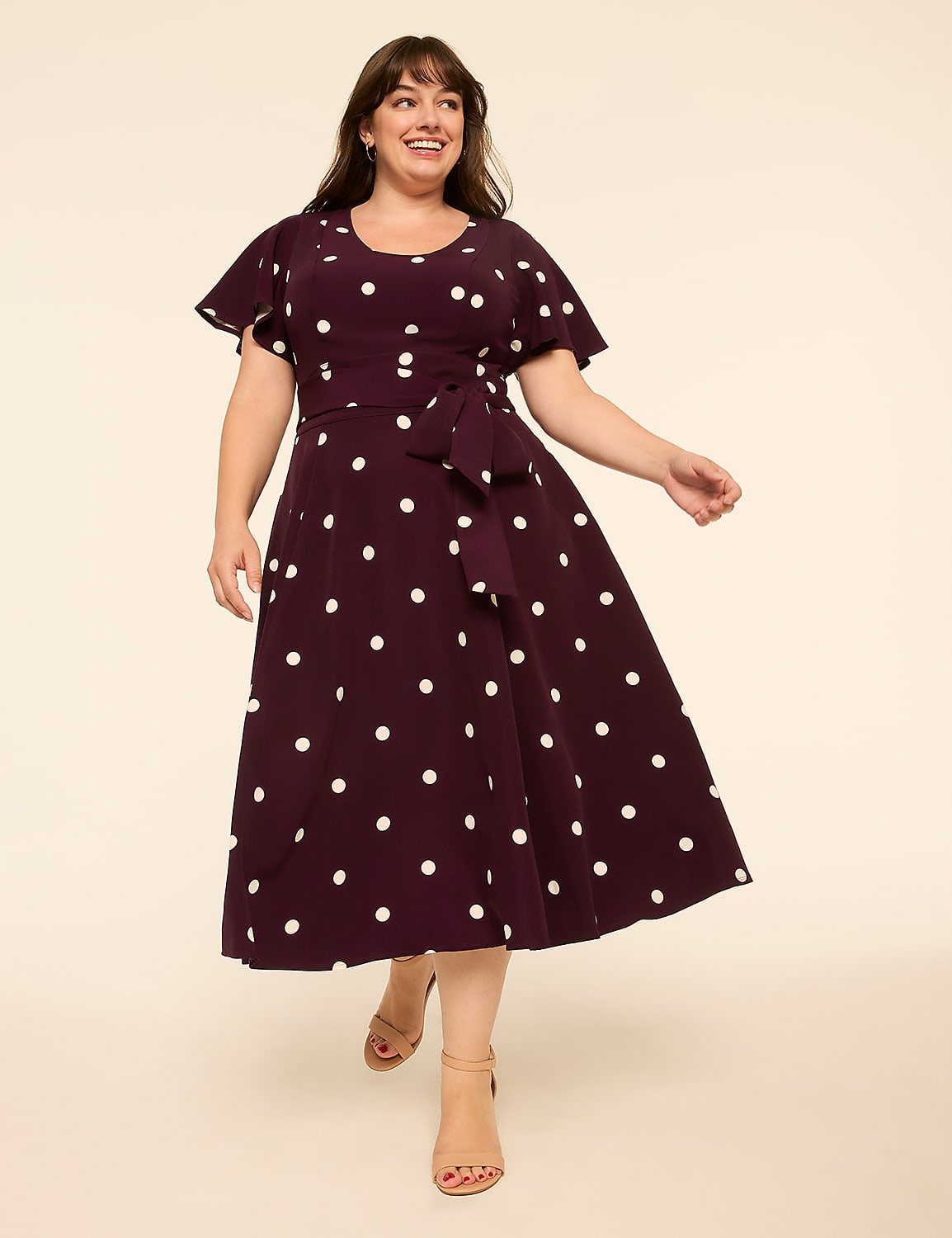 A smiling woman in a polka-dot dress walks forward against a plain, light-colored background. Product Image 1
