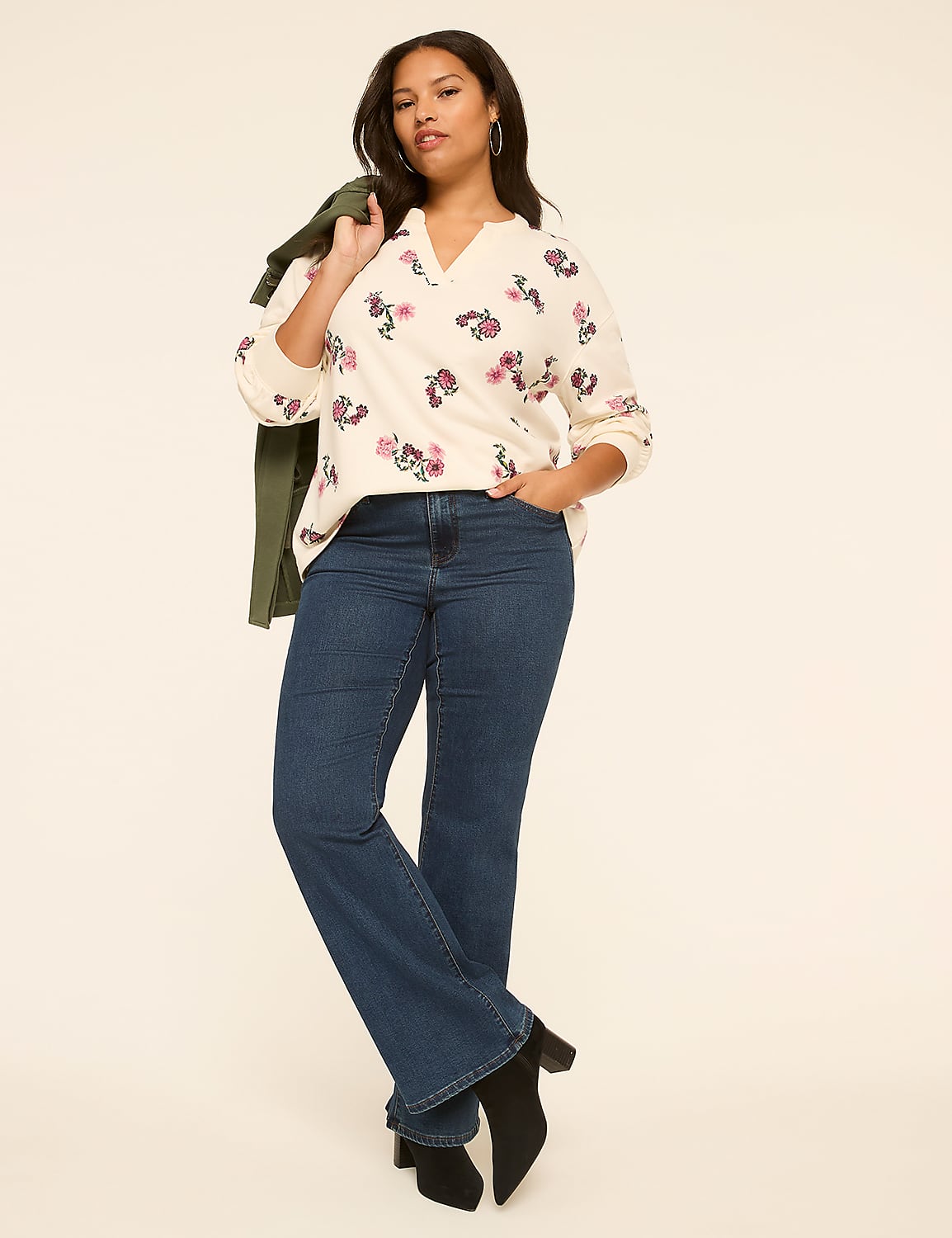 A woman in a floral sweater and flared jeans stands against a plain, light-colored background, with a green jacket draped over her shoulder. Product Image 3