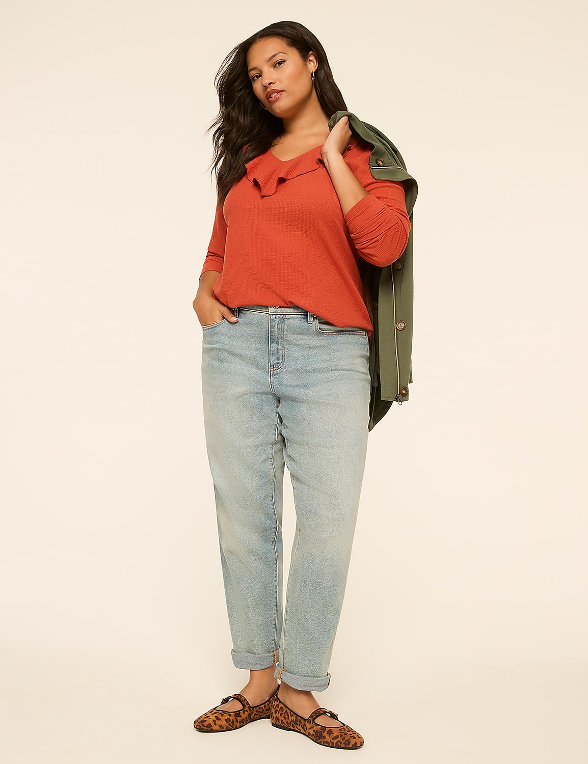 A woman in a rust-colored top, light-wash jeans, and leopard print flats stands against a plain, light-colored background with a green jacket draped over her shoulder. Product Image 3