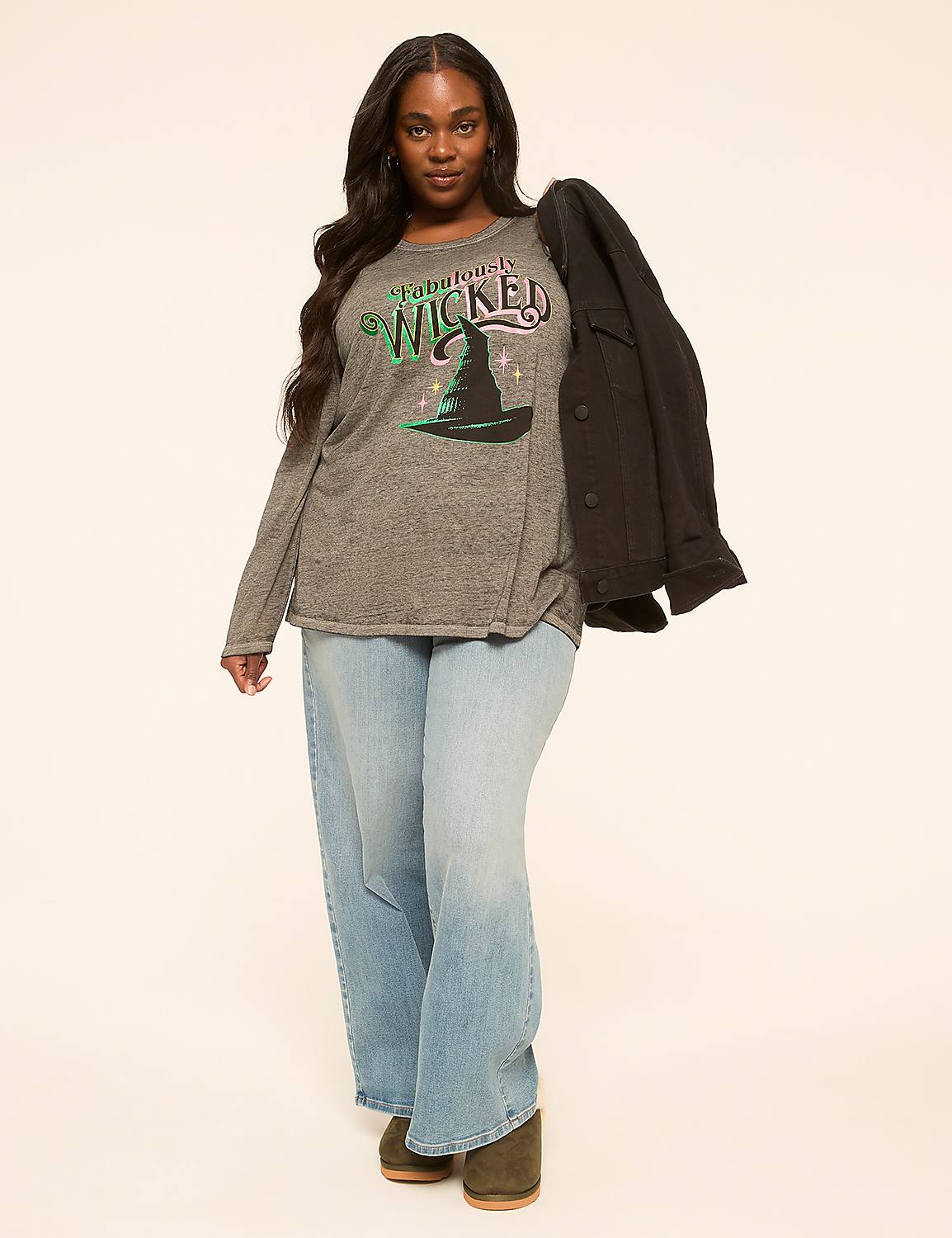 A woman in a grey graphic t-shirt and light wash jeans stands against a plain, light-colored background, with a black jacket draped over her shoulder. Product Image 3