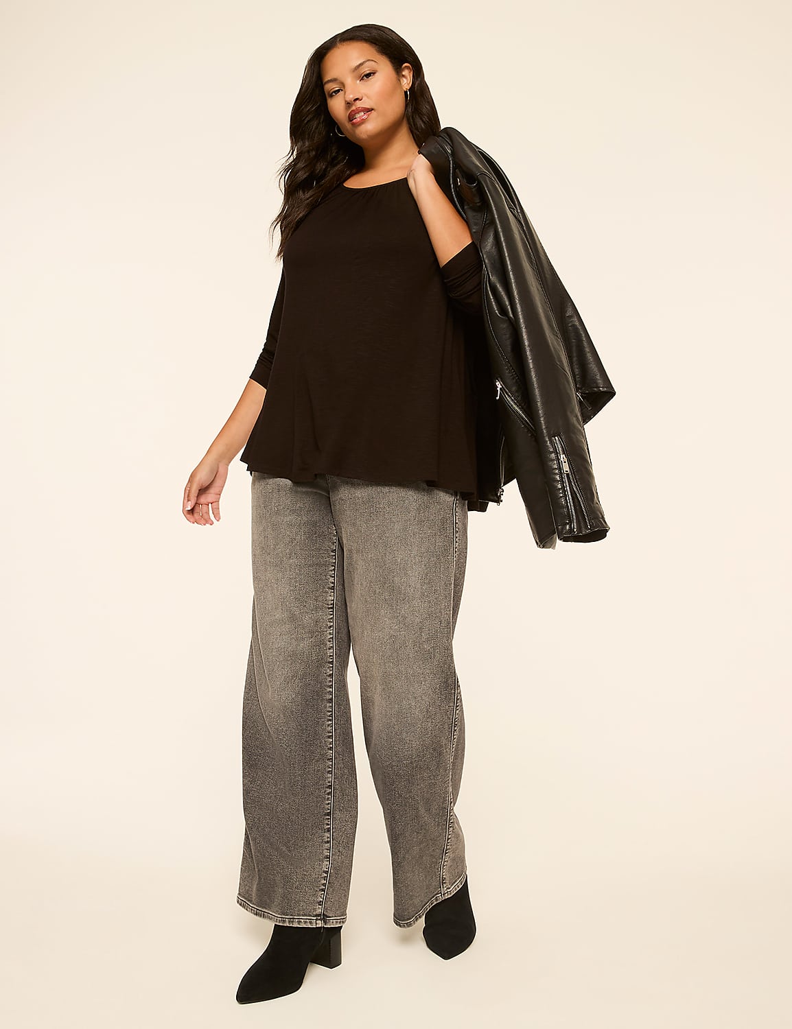 A woman in a black top, grey wide-leg jeans, and black boots stands against a plain, light-colored background with a black leather jacket draped over her shoulder. Product Image 3
