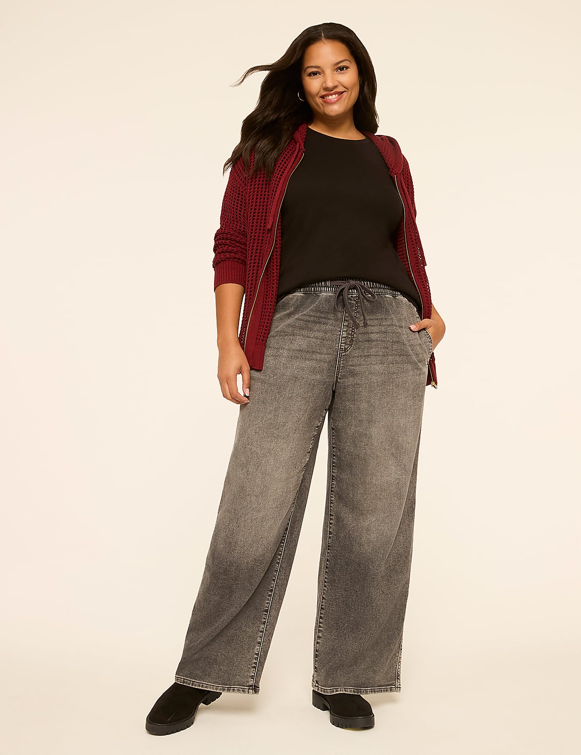 A smiling woman in a maroon zip-up sweater and grey wide-leg jeans stands against a plain cream background. Product Image 3