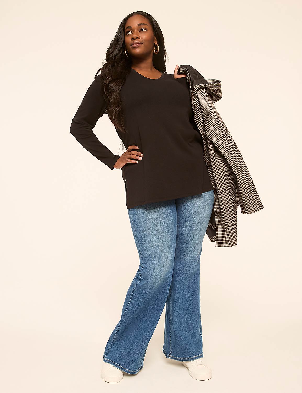 A woman in a black top and flared jeans stands against a plain, light-colored background, with a patterned coat draped over her shoulder. Product Image 3