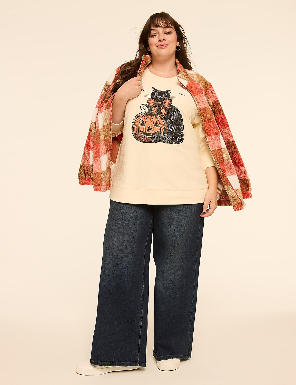 A woman in a cream-colored sweatshirt with a black cat and pumpkin graphic, dark wide-leg jeans, and a plaid jacket draped over her shoulders stands against a plain, light-colored background. Product Image 3