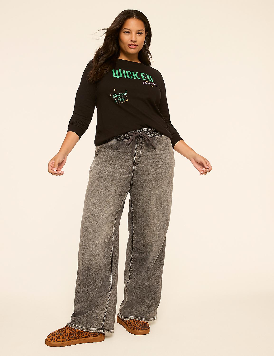 A woman in a black "Wicked" t-shirt and grey wide-leg jeans stands against a plain, light-colored background. Product Image 3