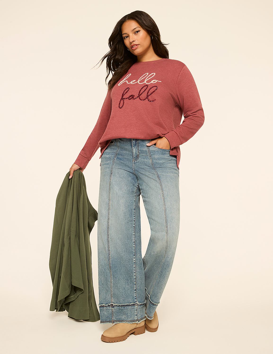 A woman in a "hello fall" sweatshirt and wide-leg jeans stands against a plain, light-colored background, holding a green jacket. Product Image 3