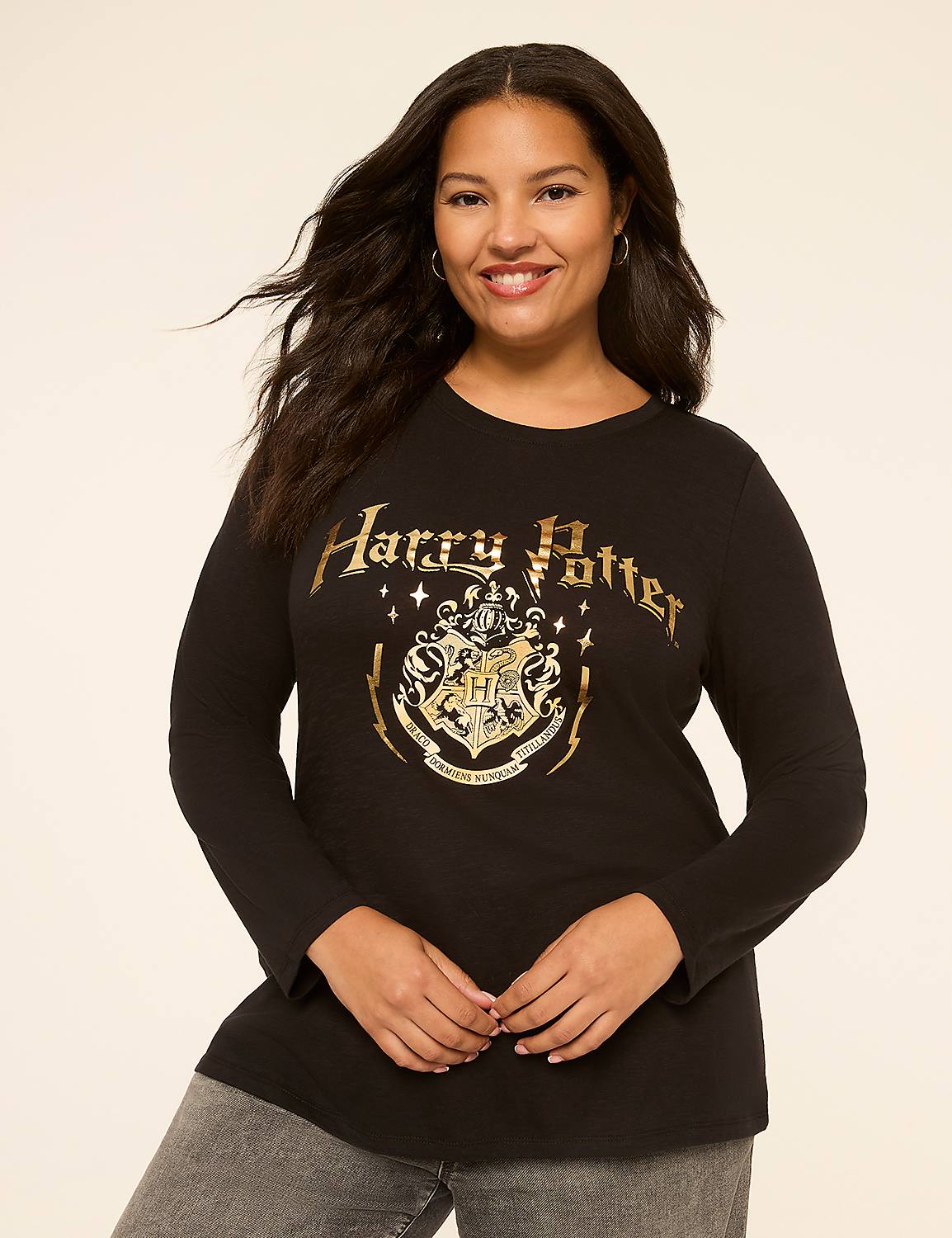A smiling woman with dark, wavy hair wears a black Harry Potter t-shirt with a gold crest and text, standing against a plain, light-colored background. Product Image 1