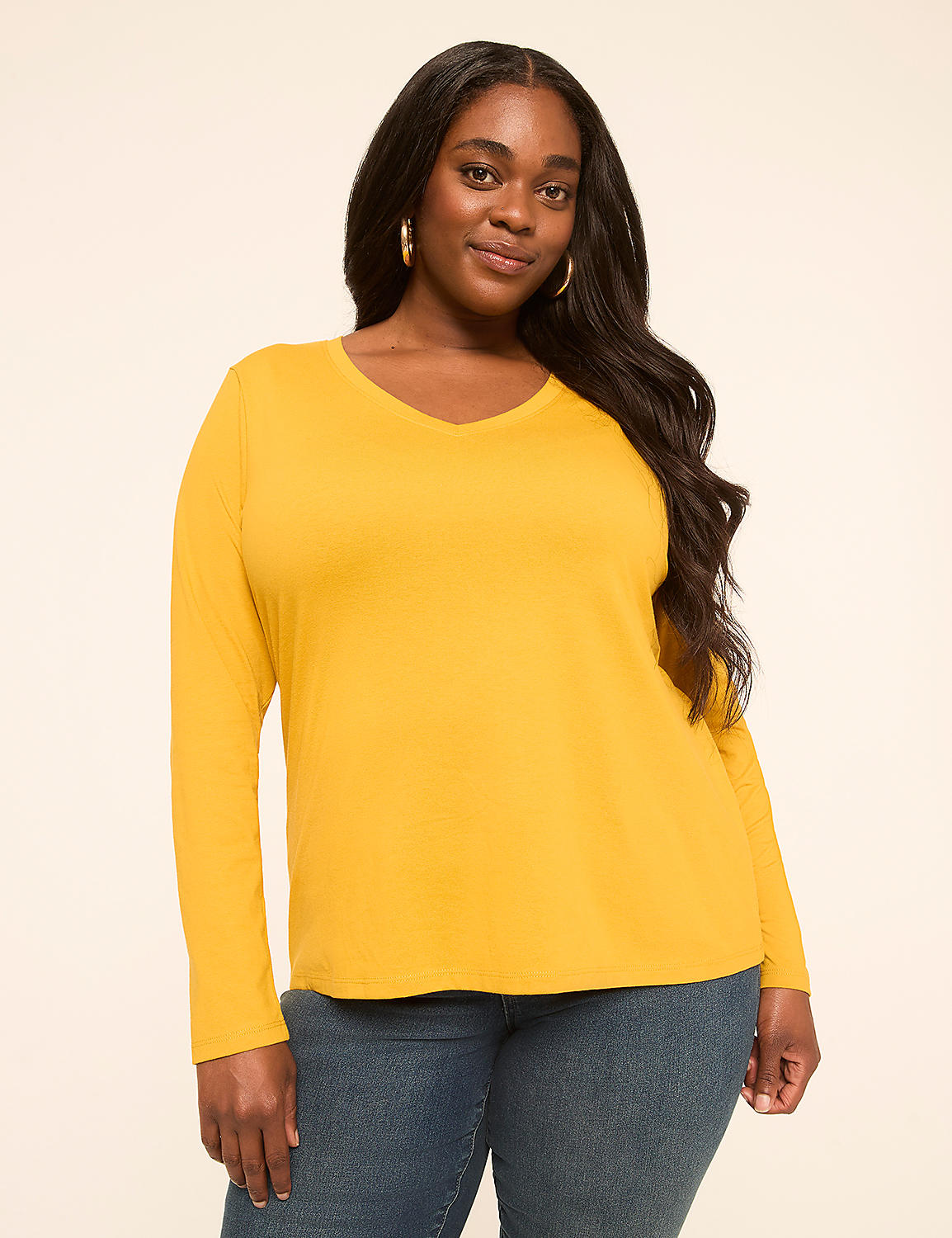 A woman with long dark hair and hoop earrings, wearing a mustard yellow long-sleeved v-neck shirt and blue jeans, stands against a plain, light-colored background. Product Image 1