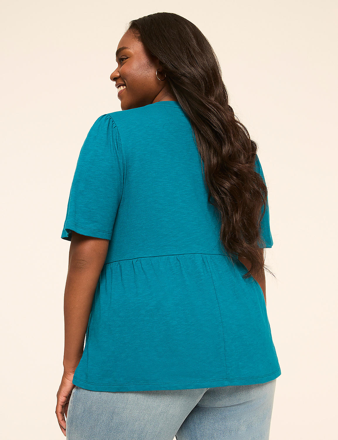 A woman in a teal top and jeans smiles over her shoulder against a plain, light-colored background. Product Image 2